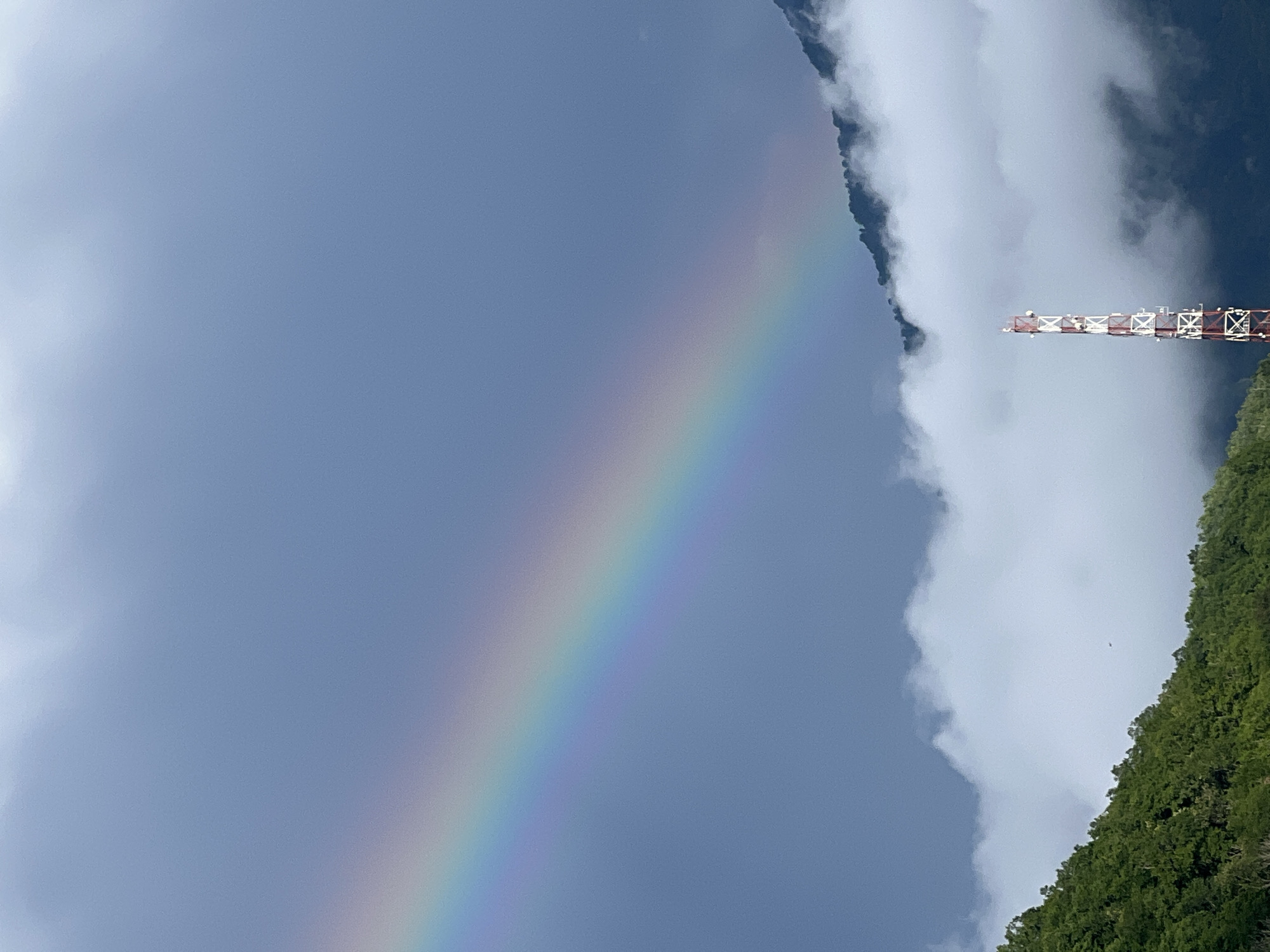 The image captures a wide-angle view of a mountainous landscape, likely in Krasnaya Polyana, Russia, under a cloudy sky with a visible rainbow. In the foreground, a lush green forested slope occupies the lower left quadrant. A tall, white and red cell tower stands prominently in the lower right quadrant, rising above a layer of white clouds that partially obscure the dark, forested mountains behind it. A distinct rainbow arcs across the upper left portion of the sky, its colors ranging from violet to red. The sky itself is a muted blue-gray, with darker clouds further obscuring the upper reaches of the mountains. The overall impression is of a natural landscape after a rain shower, with the rainbow adding a touch of color and hope to the overcast conditions.