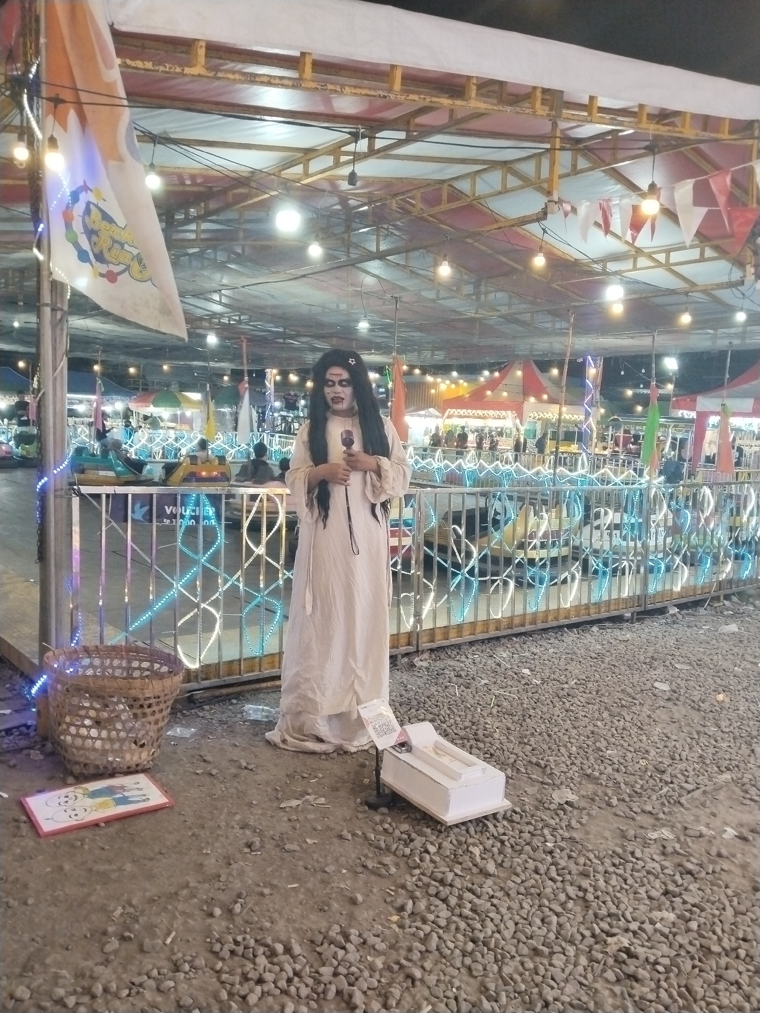 The image captures a night scene at an outdoor fairground or amusement park in Sragen, Indonesia. The central figure is a person dressed in a ghost or horror-themed costume, characterized by a long white gown, long dark hair, and white face paint with dark eye and mouth makeup, and a black star drawn on their forehead. They are holding a microphone, suggesting they are a performer or entertainer.
To the left of the performer, a wicker basket sits on the gravel ground, next to a framed picture of two cartoon characters, possibly resembling Doraemon and Nobita or similar style. To the performer's right, a small white box, likely a donation box or prop, rests on a stand with a visible QR code.
The background reveals a lively night market atmosphere with numerous string lights, overhead lights, and decorative flags. A large white flag hanging from a structure displays partial text that appears to be "Beek Ria" and a colorful logo. Behind the performer, a metal fence adorned with blue and white LED strip lights encloses an area with bumper cars, some of which are occupied by people. Additional tents and stalls with various lighting are visible in the distance. The ground throughout the scene is covered in gravel and dirt. Visible text on a sign behind the fence includes "VOUCHER" followed by some Indonesian words. The overall setting is brightly lit, indicative of nighttime activity.