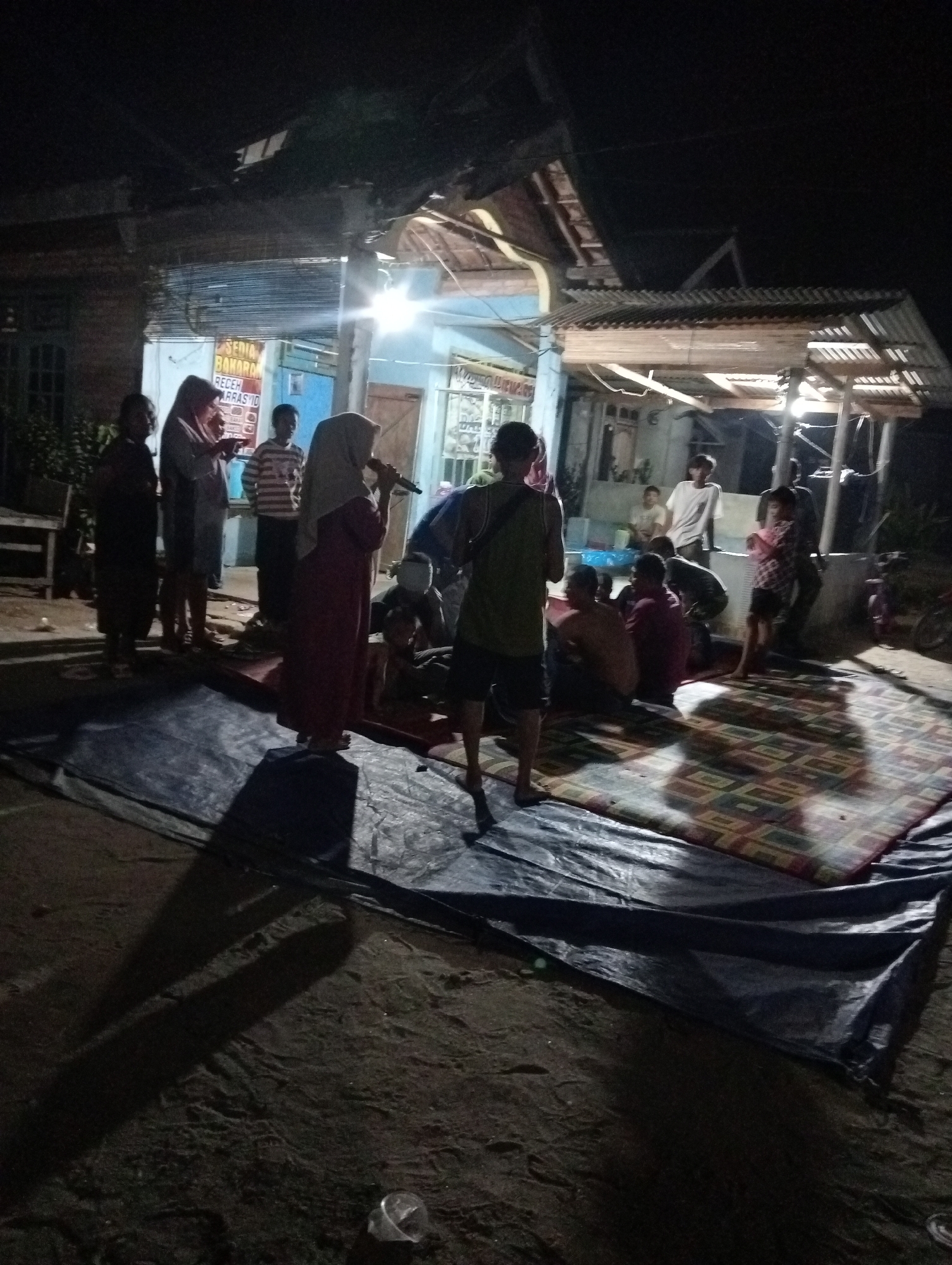 The image depicts an outdoor gathering at night in Rasuan Baru, Indonesia. The scene is illuminated by artificial lights from a building that appears to be a roadside stall or shop. Several individuals are present, with some standing and others sitting on mats laid out on the ground. One person, a woman wearing a headscarf, is holding a microphone, suggesting a performance or speech is taking place. Another individual, a man in a green tank top and shorts, stands facing away from the camera. The ground is dirt, and a large, dark tarp is spread out on the left side of the frame. A patterned mat is also visible on the right, where some people are seated. The building behind the gathering has a corrugated metal roof and visible windows. The overall atmosphere appears casual and communal.