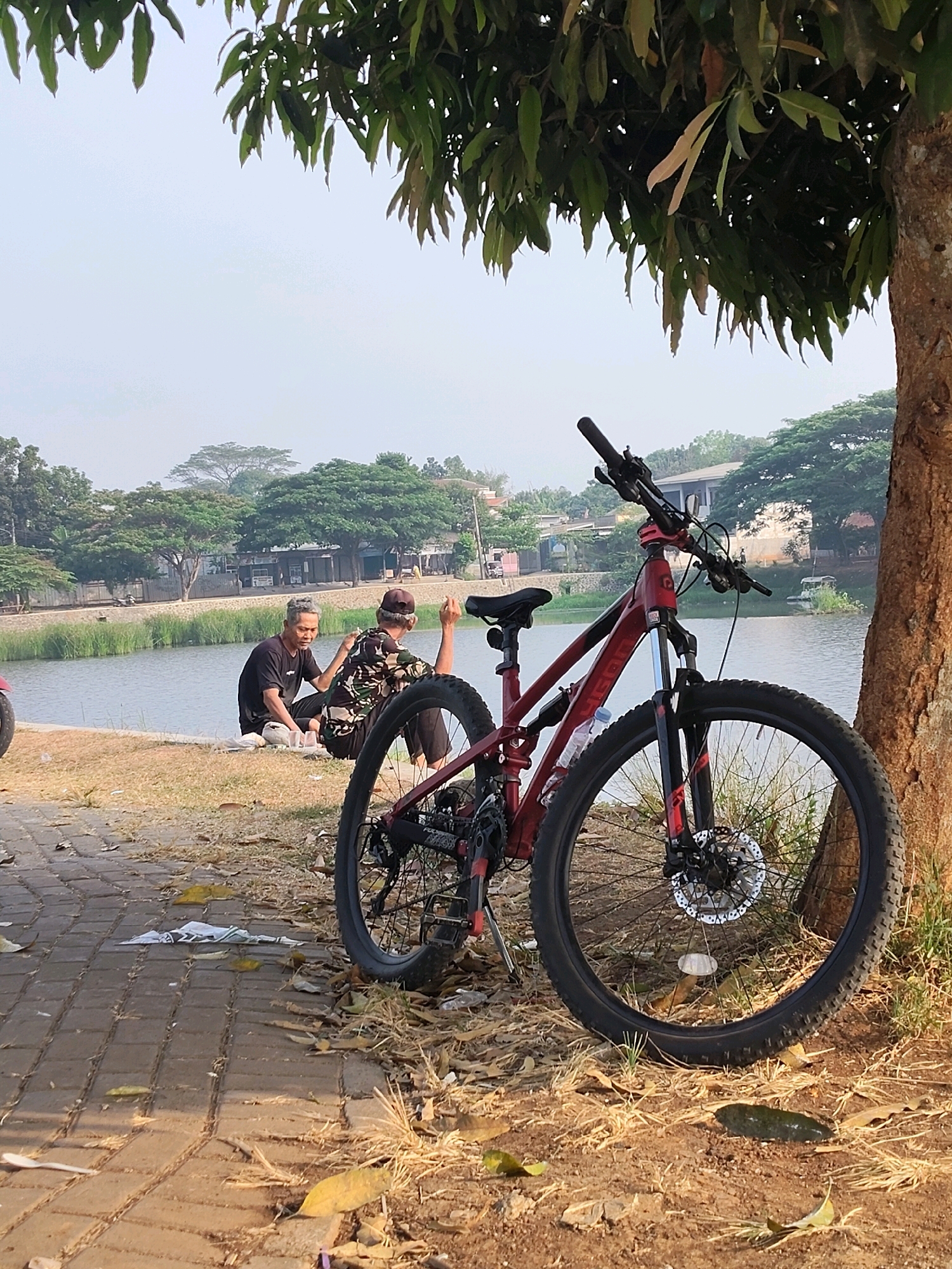In Cileduk, Indonesia, two men are seated on the grassy bank of a body of water, engaged in conversation. One man, wearing a black t-shirt, gestures with his hands while the other, in a camouflage patterned shirt and a brown cap, looks in the same direction. A red bicycle is parked nearby, leaning against a tree trunk. The ground is covered with a mix of dirt, dry leaves, and a paved walkway. In the background, lush green trees and buildings line the opposite side of the water. The sky is hazy, suggesting a warm, possibly humid day.