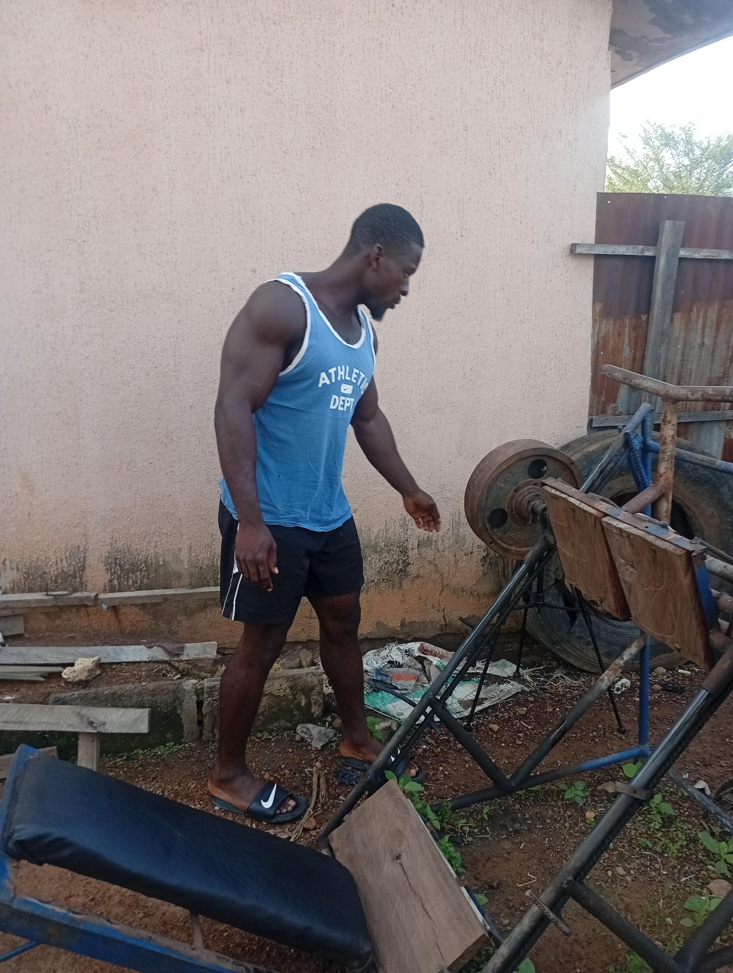 A muscular Black man is depicted standing in what appears to be an informal outdoor gym. He wears a light blue tank top with "ATHLETE DEPT" printed on the chest, black shorts with white side stripes, and black Nike slide sandals. His hair is short, and he has a beard. He is positioned in the center-left of the frame, looking down and slightly to his right with a focused expression, his right hand lightly extended.
To his right stands a rustic, possibly homemade, leg press machine. It is constructed from metal bars, featuring a wooden footplate and a large, heavily rusted circular weight plate. In the foreground on the left, part of a workout bench with a black upholstered pad is visible. The ground is uneven, a mix of dirt, small rocks, and patches of green weeds.
The background features a light beige or tan-colored plastered wall. To the right, a weathered, rusty corrugated metal fence is visible, with green foliage and tree branches extending above it, suggesting an open-air environment. Various discarded items, including old tires and wooden planks, are scattered around the equipment.
The scene is illuminated by bright daylight, indicating a clear morning or afternoon. The man appears to be either preparing for an exercise, observing the equipment, or taking a short break. The setting, with its improvised equipment and outdoor nature, suggests a functional training space in a location such as Akwanga, Nigeria.