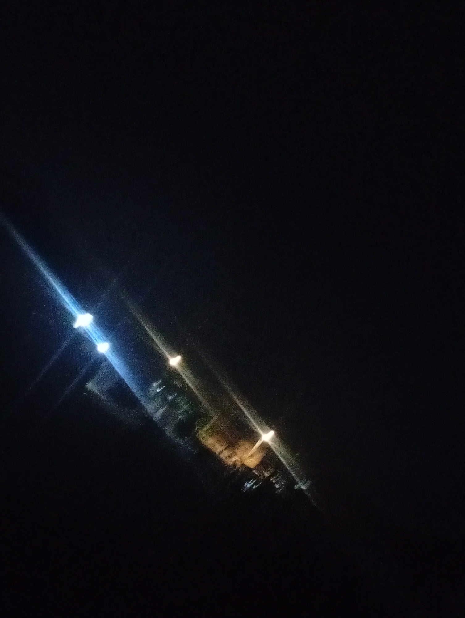 The image, taken in Bukuru, Nigeria, depicts a nighttime scene with artificial lights illuminating a dark environment. In the center of the image, several lights are visible, appearing as streaks of white and yellow due to lens flare and low light conditions. These lights seem to originate from a structure, possibly a building or a cluster of vehicles, indicated by indistinct shapes and shadows beneath them. The overall impression is that of an urban or semi-urban setting at night, with limited visibility and a focus on artificial light sources. The background is entirely black, suggesting a lack of ambient light and potentially a distant perspective.