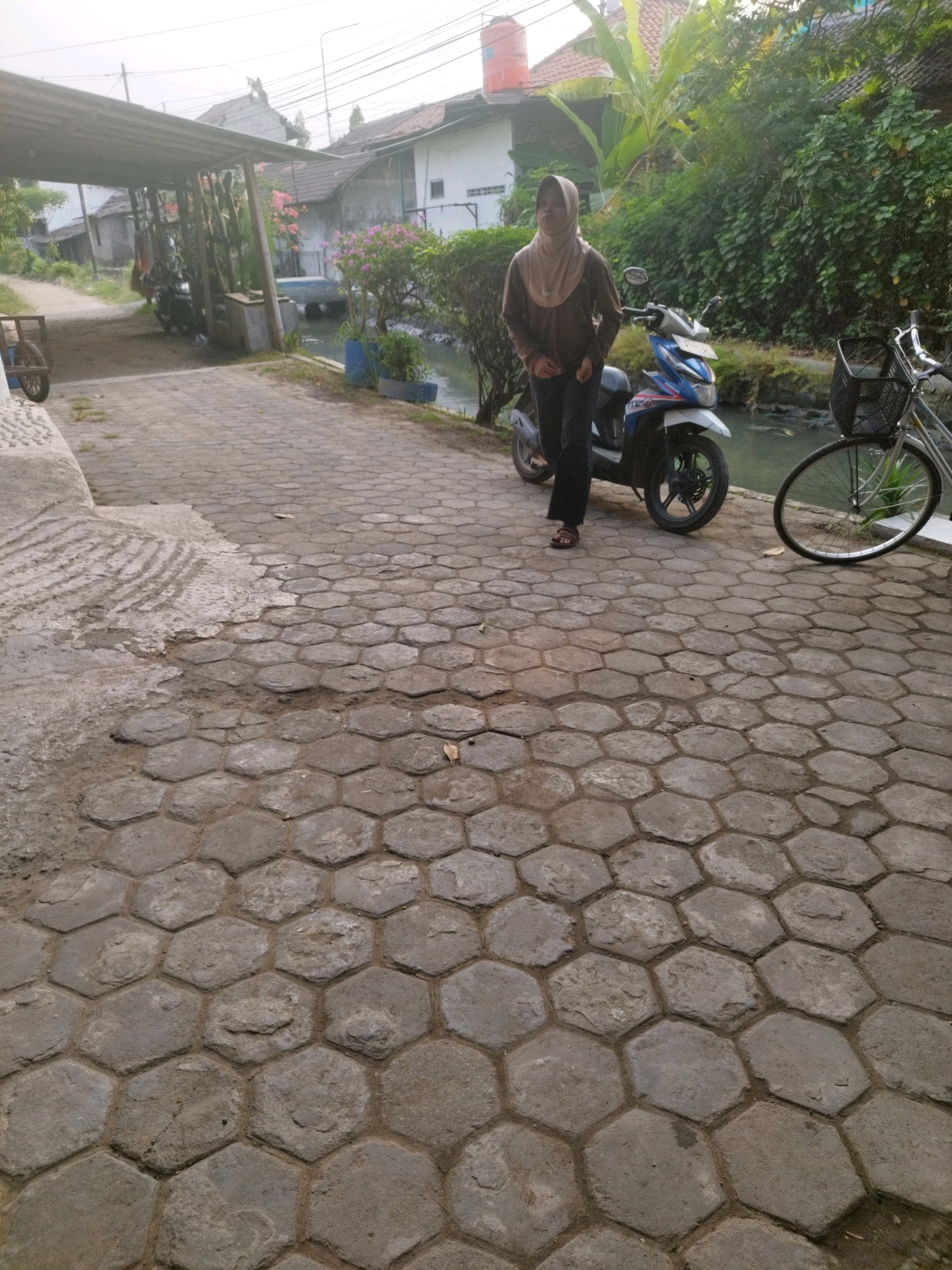 In Patebon, Indonesia, a young woman wearing a light brown hijab and a dark brown long-sleeved shirt stands on a hexagonal-patterned paved walkway. She is looking to her left. To her right, a blue and white scooter is parked. Further to the right, a bicycle with a basket is visible. Behind the woman and scooter, a small canal or body of water flows, bordered by greenery and houses. A covered structure, possibly a carport, is visible on the left side of the image, with a motorcycle parked underneath. The overall scene depicts a residential area with a mix of paved surfaces, natural elements, and common modes of transportation.
