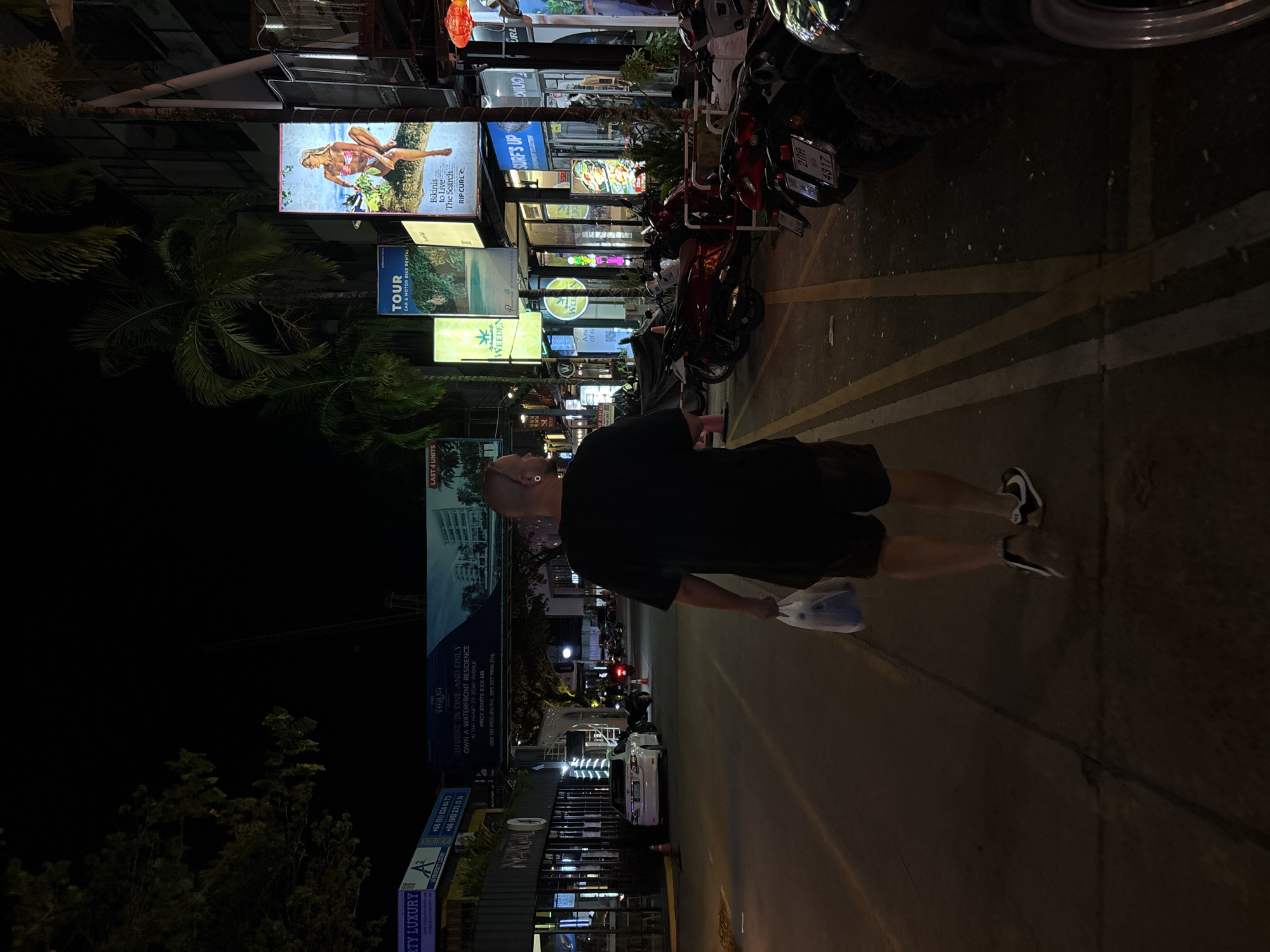 A person walks down a street at night in Choeng Thale, Thailand. The street is lined with various shops and signs, many of them illuminated. The person is wearing dark clothing and holding a plastic bag. The scene appears to be a commercial area with shops selling services like car and motorbike rentals, as well as advertisements for residences. One prominent sign features an image of a woman in a bikini and the Rip Curl logo, indicating a surfing or beachwear brand. Other signs mention "Tour," "Surf's Up," and real estate advertisements with Thai text and phone numbers. The presence of parked motorcycles suggests a busy area, possibly a tourist destination. The time of day is clearly night due to the darkness and the illuminated signs. The weather is not discernible from the image.