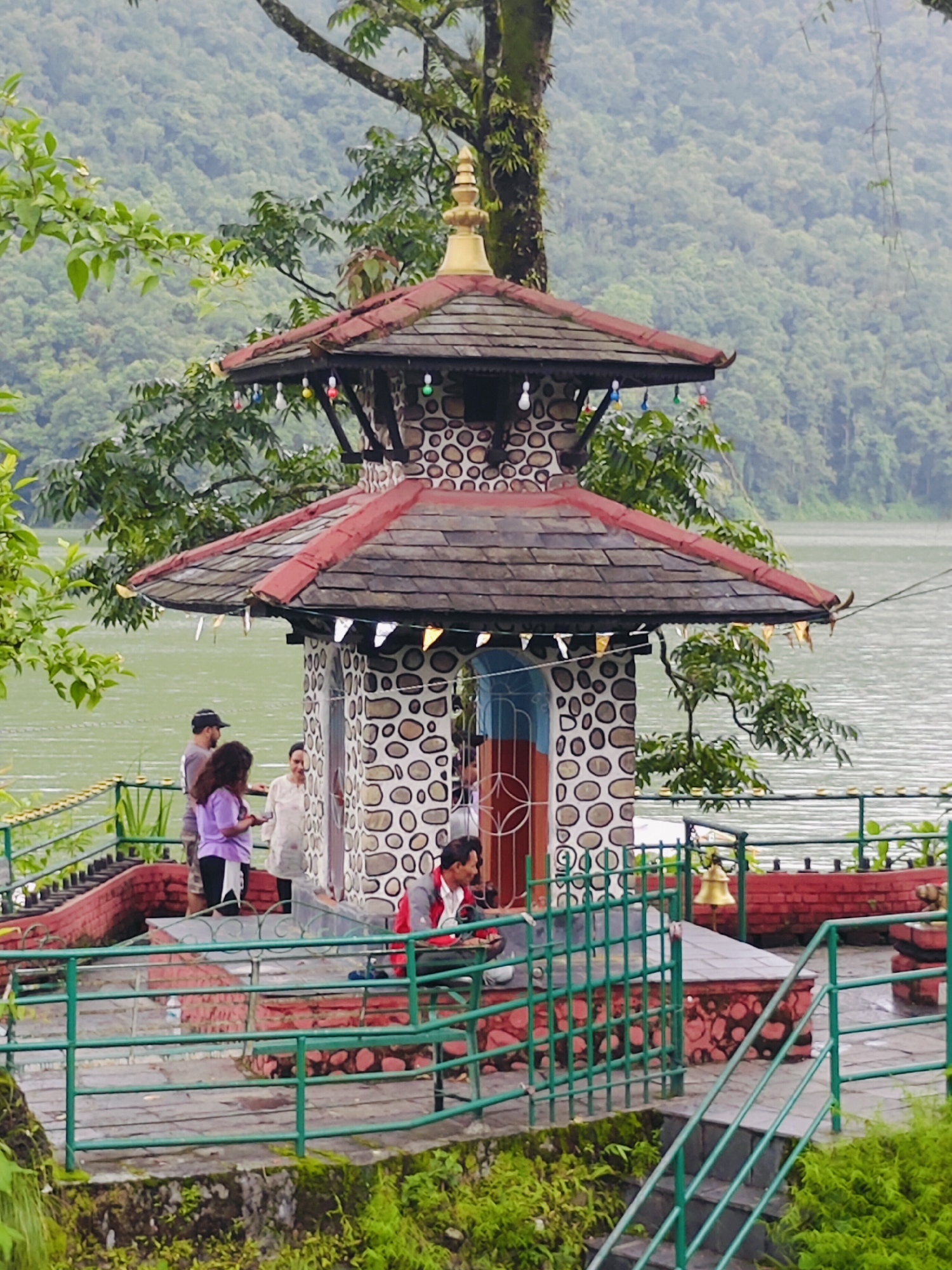 A two-tiered pagoda-style building with a red roof and stone and white circular patterned walls stands on a raised platform beside a lake. Several people are present: two women and a man stand near the entrance of the building on the left, one woman in a purple jacket and another in a white top, and a man with a dark cap. On the right, a man wearing a red jacket sits on a bench. A golden bell hangs on a post to the right of the building. The area is enclosed by a green metal railing. The background shows a lush, green, forested hillside under a cloudy sky. The scene is likely at the Barahi Temple on Phewa Lake in Pokhara, Nepal.