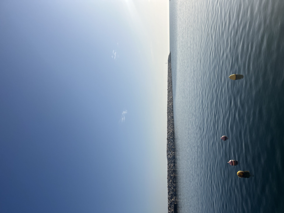 The image shows a view of the calm sea from a harbor, with a rock breakwater in the foreground. The breakwater is made of large, irregularly shaped rocks and extends across the entire width of the image. The sea is a deep blue color, with a slight ripple. There are four buoys in the water, two pink and two yellow. The sky is a clear blue, with a few wispy white clouds. This view was captured at 40.625 latitude and 24.578 longitude, which is the location of the port of Limenaria on Thasos island, Greece. There are no people visible.