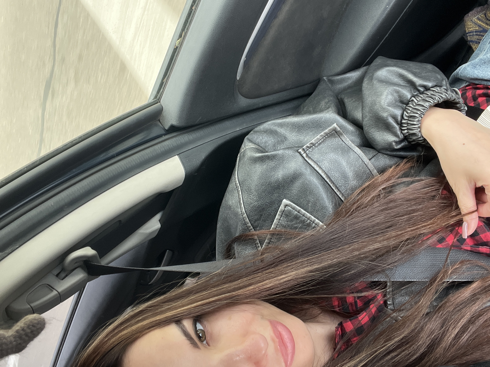 The image depicts a person sitting inside a vehicle. The person is wearing a red and black plaid shirt and a dark gray leather jacket with a ribbed cuff. A seatbelt is fastened across the person's body. The interior of the vehicle includes a gray trim and dark gray door panel. Outside the window is an indistinct, light-colored surface. The city location is unknown.