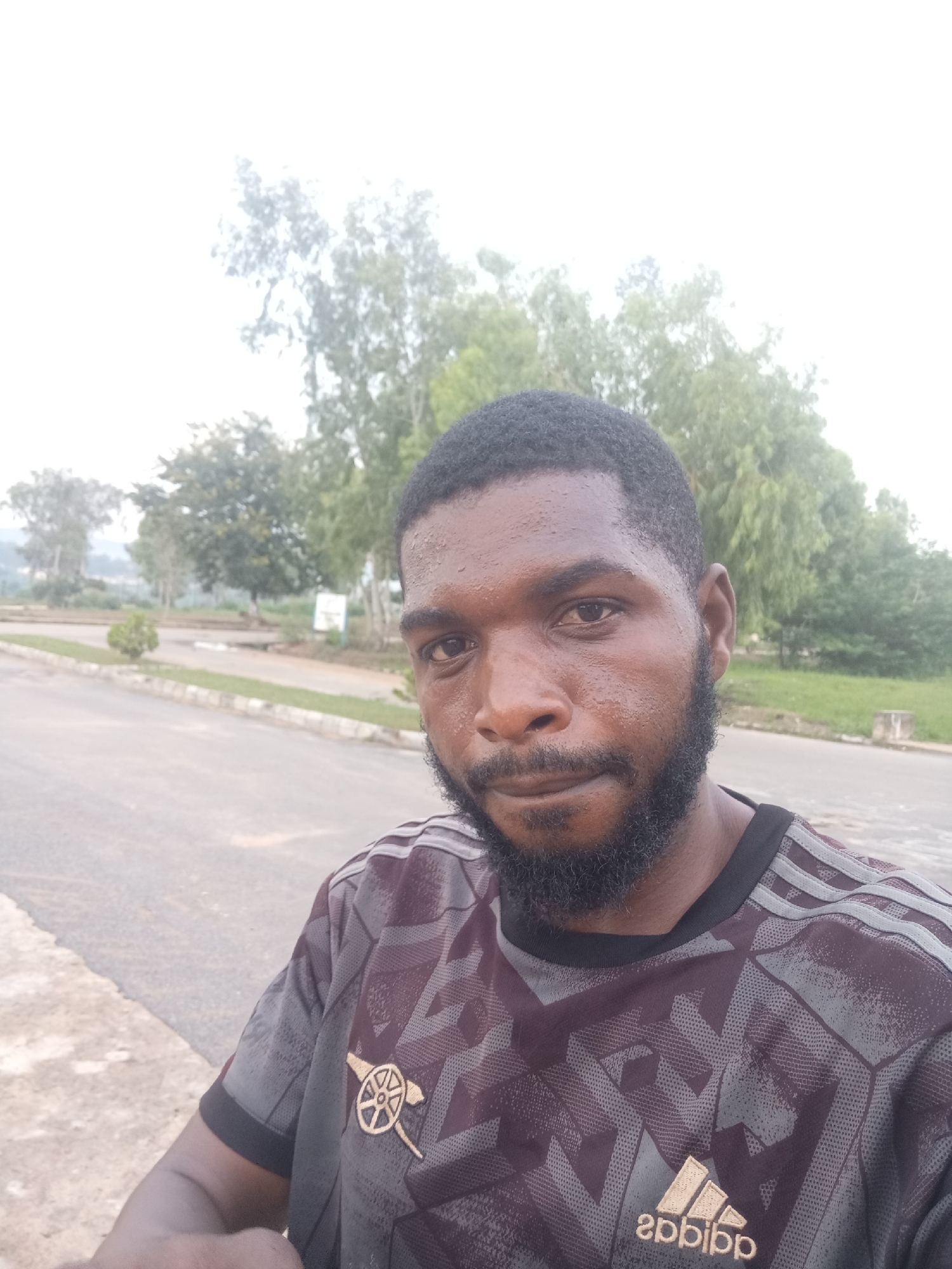 A man with dark skin, short black hair, and a beard is taking a selfie outdoors. He is wearing a dark t-shirt with a patterned design and logos. The background shows a paved road, a grassy area with a small tree, and more trees in the distance. The sky is overcast, suggesting an outdoor setting possibly in Jos, Nigeria, given the overall environment and vegetation.