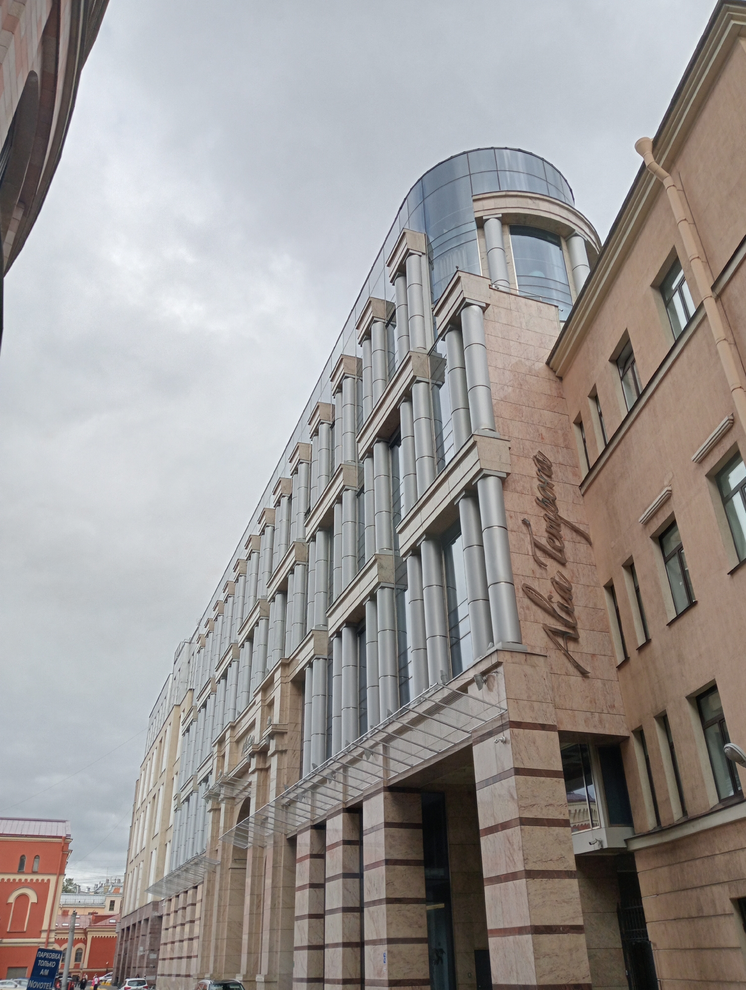 The image captures a low-angle view of a modern building facade in Saint Petersburg, Russia, under a cloudy sky. The building is characterized by its polished stone cladding in shades of beige and brown, with distinct horizontal bands of darker marble. It features a repetitive design of tall, slender cylindrical columns that run vertically along its facade, interspersed with large glass windows. A curved glass section is visible at the top of the building. A metallic awning with a transparent roof extends over the entrance. To the right of this modern structure, an older, light-peach colored building with smaller windows is visible, displaying architectural elements such as window casings and a downspout. The inscription "Alia Tempus" is prominently displayed in elegant, cursive script on the facade of the modern building. The scene appears to be a commercial or office building, situated in an urban environment, with glimpses of other buildings and elements of street infrastructure further down the street.