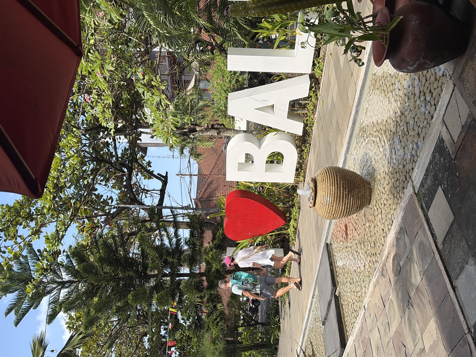 The image depicts a sunny outdoor scene in Kuta, Indonesia, with the iconic "I Bali" sign as the central focus. The sign is composed of large, white, three-dimensional letters spelling "BALI," with a prominent red heart preceding them, signifying "I."
In the foreground, a shallow water feature with smooth, light-colored pebbles is visible. A rustic, ribbed ceramic pot with a small fountain spout is positioned within the water. To the right, a dark brown ceramic pot contains green foliage. The walkway in front of the sign is paved with square tiles in various shades of brown and grey.
Two individuals are walking away from the camera on the paved path. The person on the left is a male with grey hair, wearing a light blue backpack, grey shorts, and flip-flops. The person on the right is a female wearing a white dress, a pink hat, and flip-flops.
Behind the sign, lush green trees and foliage create a vibrant backdrop. In the distance, a building with a reddish-brown tiled roof and balconies is visible, along with a blue sky. Small decorative flags on strings are strung across some of the trees on the left side of the image. The overall atmosphere is tropical and inviting.