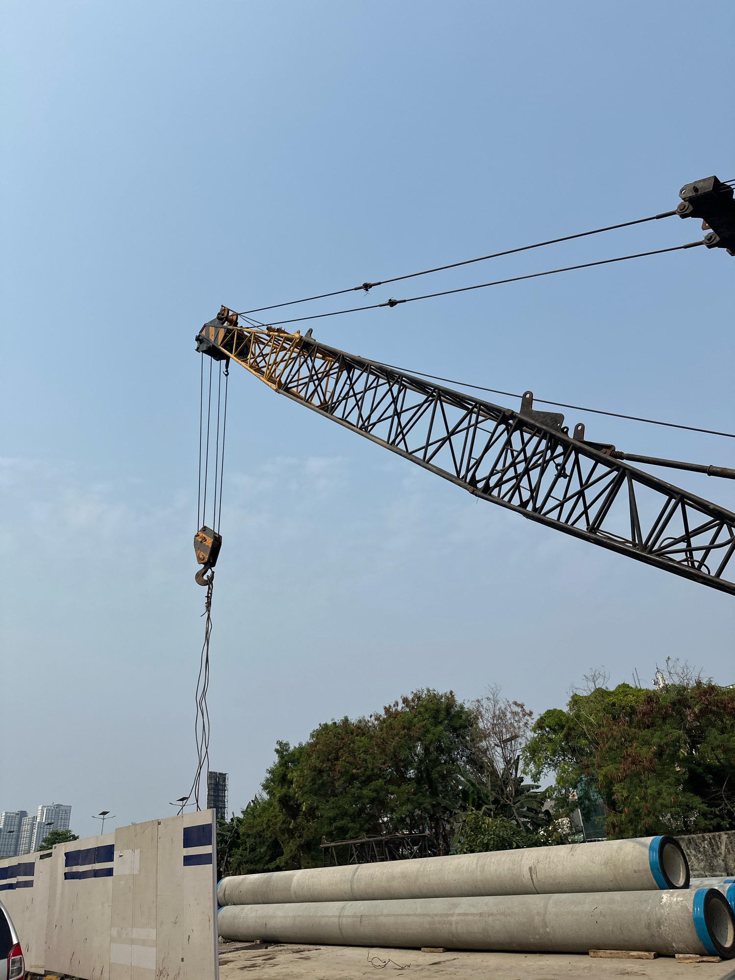 In West Jakarta, Indonesia, a construction site is visible under a clear sky. A large crane dominates the upper portion of the frame, its yellow and black lattice boom extending diagonally across the scene. A heavy hook hangs from its cables. In the foreground, large concrete pipes are stacked, suggesting ongoing infrastructure work. Behind the pipes and a construction barrier, lush green trees form a backdrop, with buildings visible in the distance, indicating an urban environment. The overall scene depicts an active construction zone within a city.