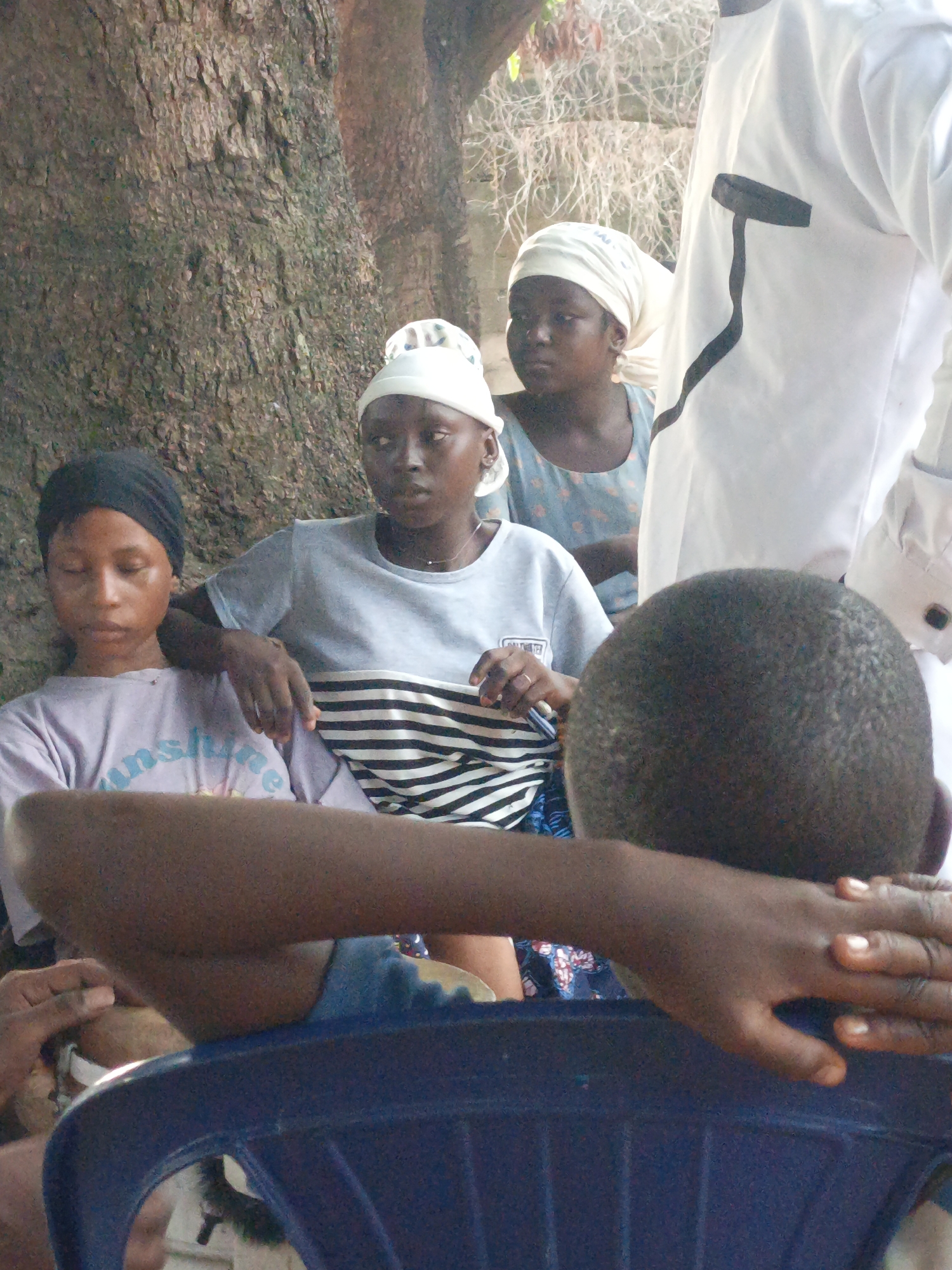 A medium shot of four people gathered outdoors in Akwanga, Nigeria. A tree trunk is visible in the background. Three women sit, two of whom are wearing white head coverings. The woman on the left wears a purple t-shirt with the word "sunshine" on it. The woman in the center wears a grey t-shirt with black and white stripes. The third woman wears a blue floral patterned dress. The fourth person, a male, stands to the right, partially visible, wearing a white garment with a black design on the chest. In the foreground, the back of a dark-haired individual seated in a blue plastic chair is visible, their arm resting on the chair's back.