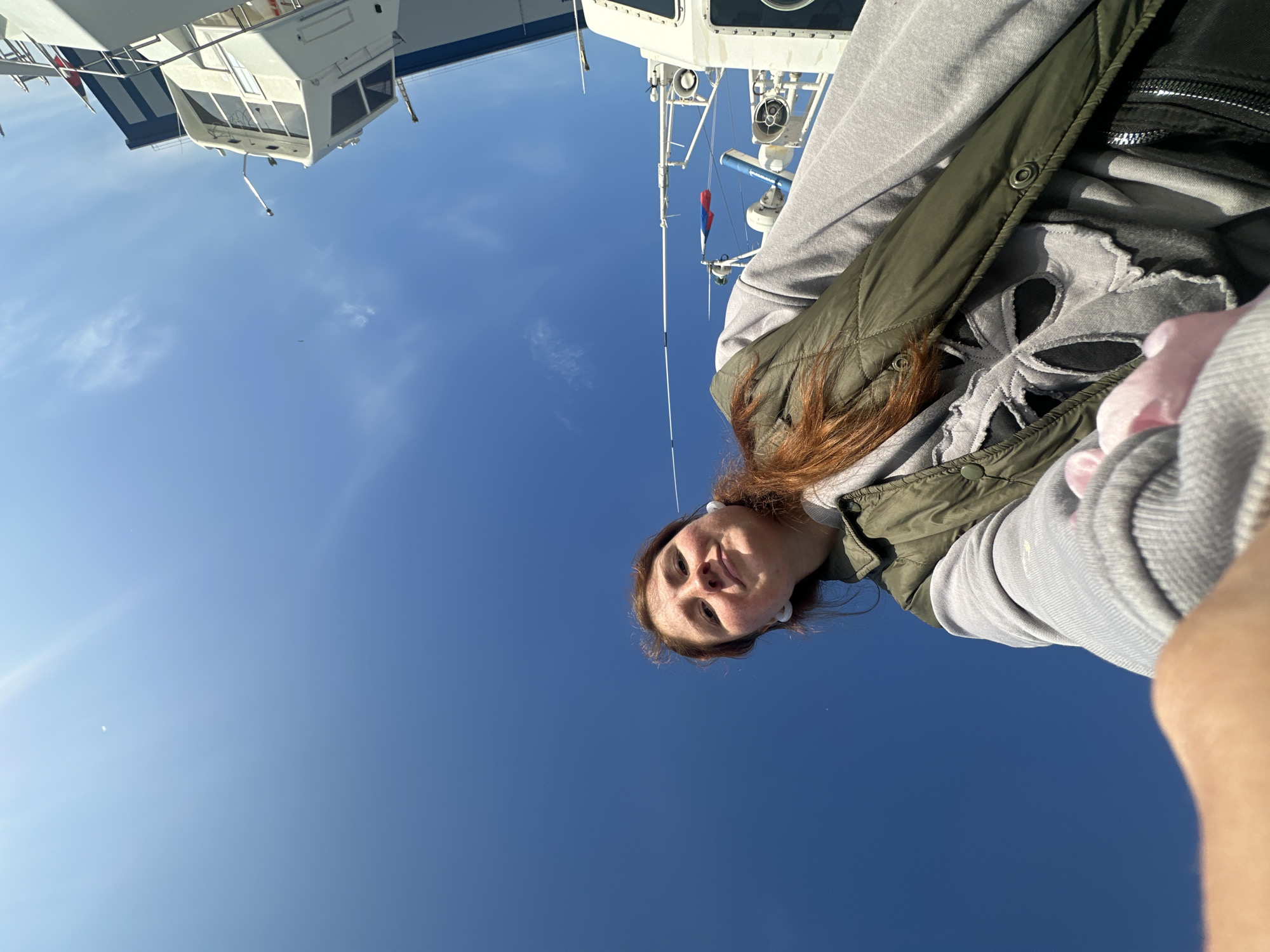 The image shows a young woman with red hair, wearing a green vest over a gray hoodie with a skull graphic. She is looking up at the camera with a smile. She is on a boat, with parts of the vessel visible in the background, including white structures and rigging against a bright blue sky. The scene appears to be in a harbor or at sea. The specific location of Petropavlovsk-Kamchatsky, Russia cannot be determined from the visual information alone.