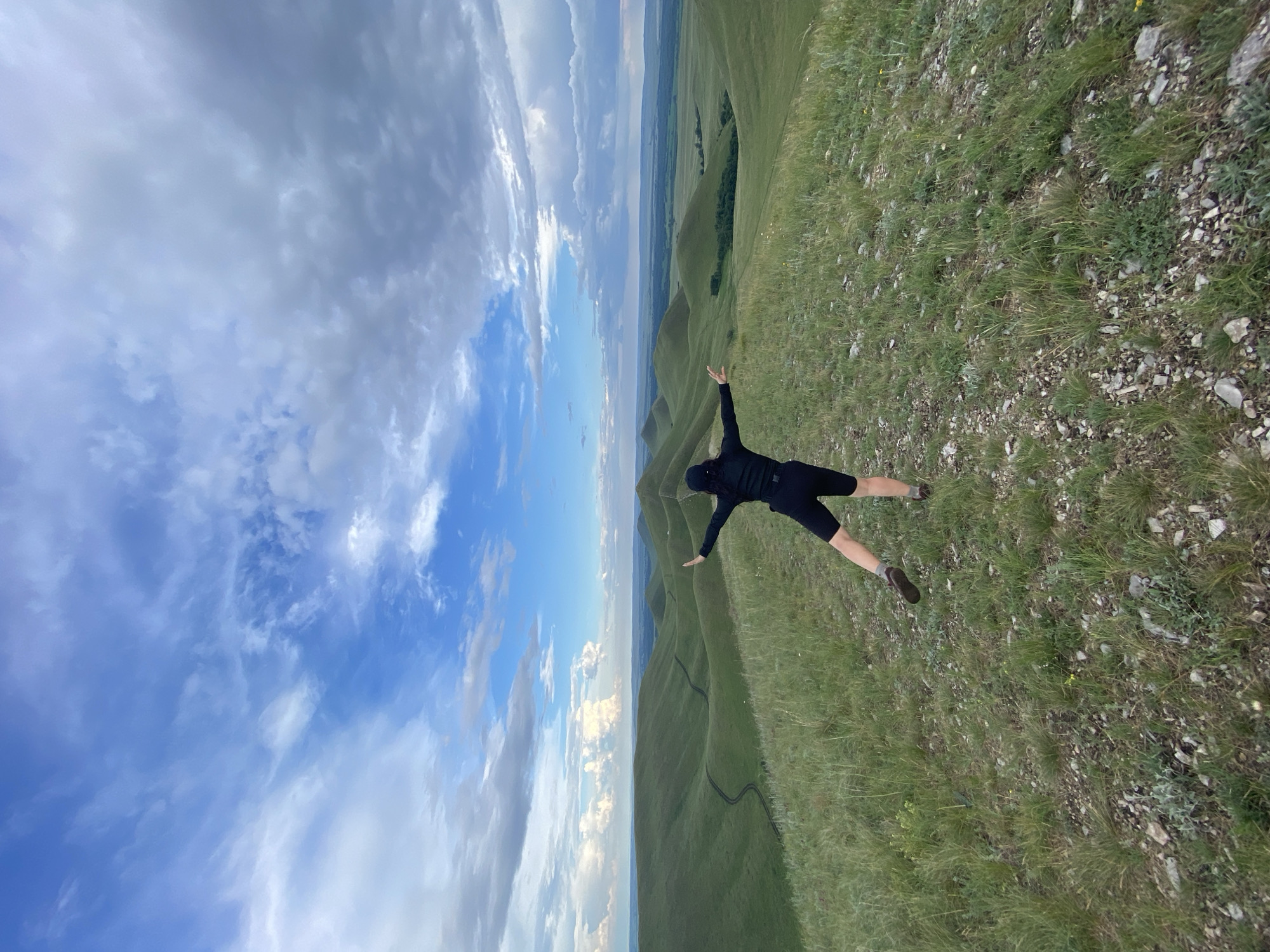 A person wearing a black cap, black long-sleeved shirt, and black shorts stands on a grassy hilltop, facing away from the camera. Their arms are outstretched to the sides, and their right leg is extended back. The person is in the process of posing or balancing. The landscape behind them features rolling green hills that recede into the distance, with a winding dirt path visible on one of the hills. The sky above is partly cloudy with a mix of blue sky and white and grey clouds. The scene appears to be a natural, undeveloped area, possibly a park or nature reserve. The location is identified as Verkhneozernoe, Russia.