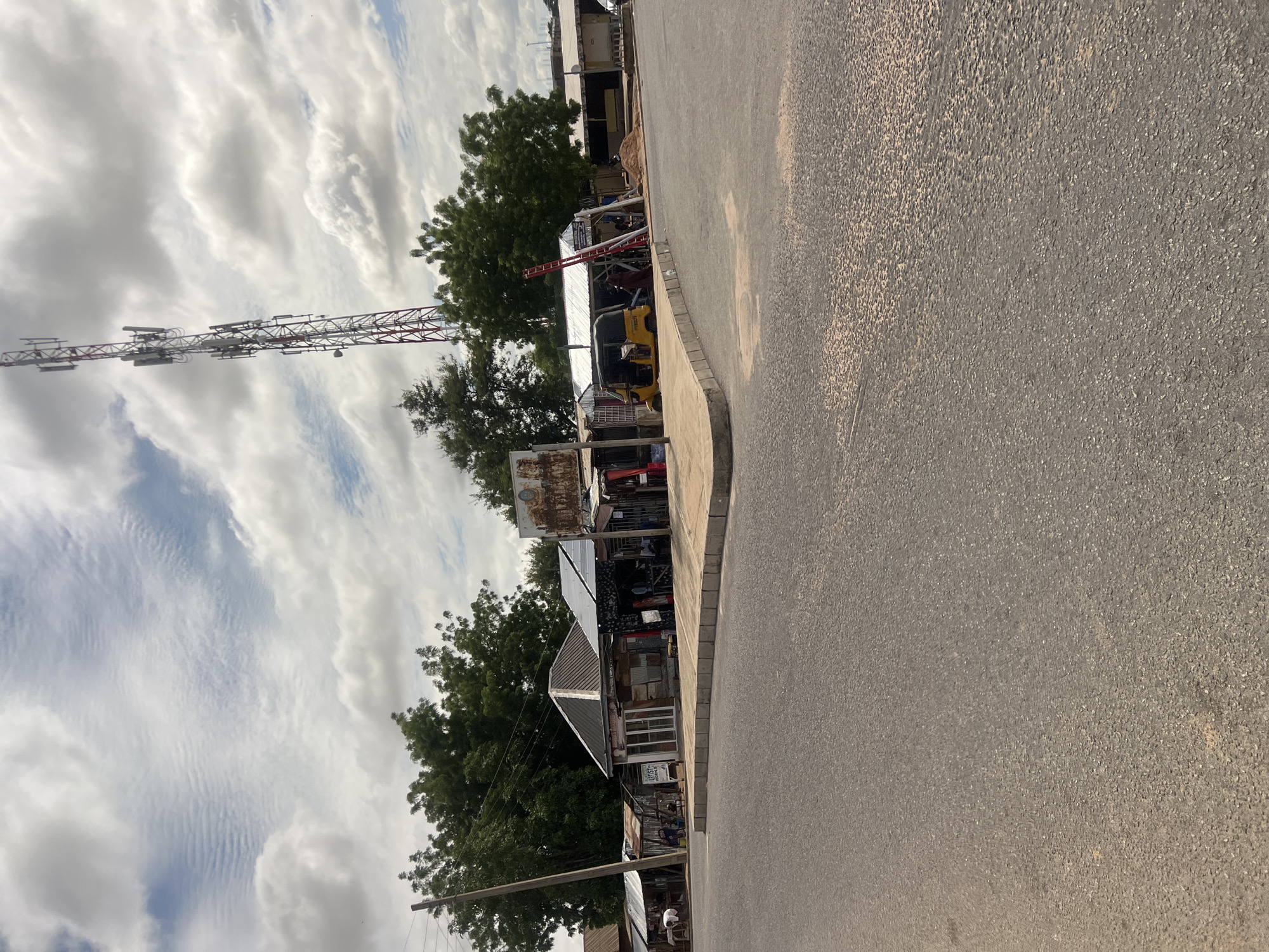 The image depicts an outdoor scene in Jalingo, Nigeria, featuring a paved asphalt road in the foreground, leading to a row of commercial structures under a partly cloudy sky.
The sky occupies the upper portion of the frame, showing a mix of white cumulus clouds and visible patches of blue. Some areas of the cloud cover exhibit a ripple-like pattern.
In the midground, a diverse collection of single-story commercial buildings or stalls lines the far side of the road. These structures predominantly feature corrugated metal roofs and varied wall materials, including wood and possibly concrete. One prominent building with a distinct hip roof and a white facade displays a partially legible sign, appearing to include the word "ARTIST." Adjacent to it, another structure has several solar panels mounted on its roof. Further to the right, a building appears to be undergoing work, with a red ladder leaning against it and a pile of sand or dirt nearby.
A yellow three-wheeled vehicle, identifiable as a Keke Napep, is parked near the shops on the right side of the road. Several individuals are present among the structures: one person in a white top is visible on the far left, another appears to be seated near the central shops, and at least one person is near the yellow tricycle. Specific actions or interactions are not clearly discernible due to distance.
A tall, red and white lattice-style cellular communication tower equipped with multiple antenna arrays dominates the right side of the image, extending into the sky. On the left, a wooden utility pole with wires is present. Lush green trees are visible behind and between the commercial structures, indicating a vegetated environment.
The foreground consists of a dark grey asphalt road with a visible aggregate texture. A raised concrete curb defines the edge of the road, curving towards the right side of the frame.