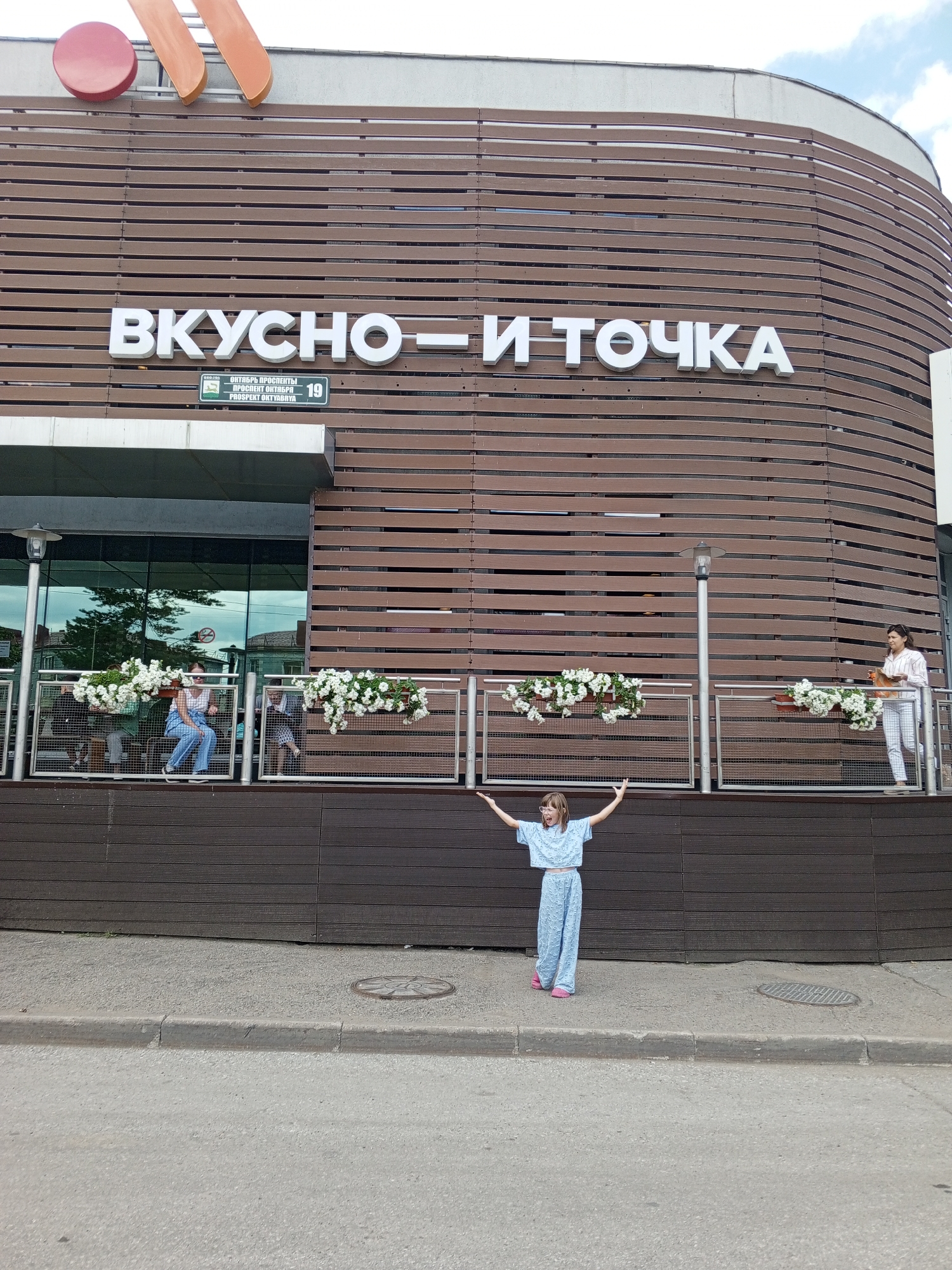 The image shows the facade of a building with the Russian text "ВКУСНО — И ТОЧКА" (Vkusno—i tochka) in large white letters. Below the text, a street sign indicates "ОКТЯБРЯ ПРОСПЕКТ" (Oktyabrya Prospekt) and the number "19". This location is in Ufa, Russia. In the foreground, a young girl in a light blue tracksuit and pink shoes stands on a paved area with her arms outstretched. She is facing the camera and appears to be looking up. Behind her, there is a wooden-paneled building with windows and a metal railing adorned with hanging flower baskets. Several people are visible on the elevated section behind the railing. The sky is partly cloudy.