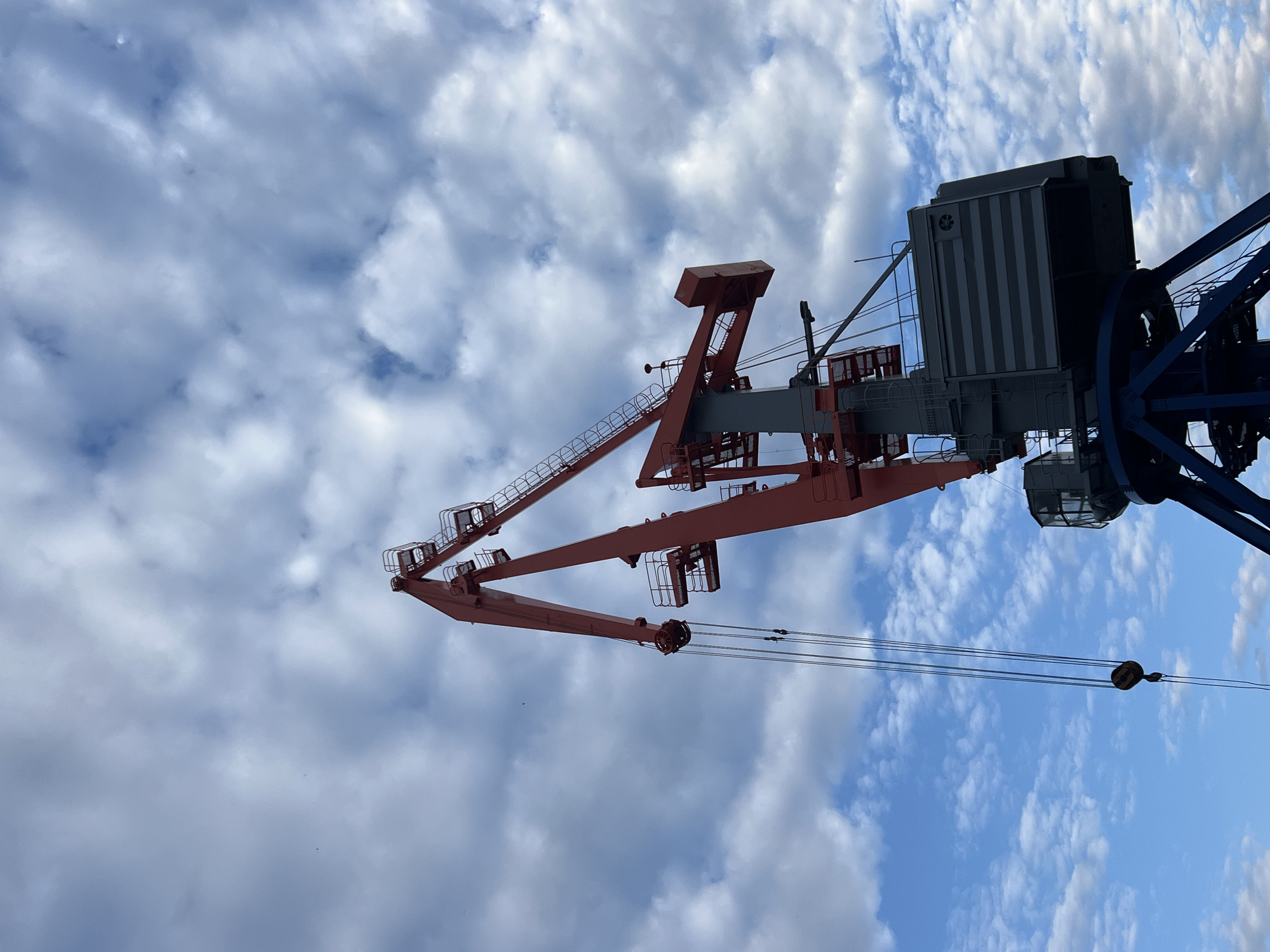 The image shows a close-up, upward-angled view of a large industrial crane against a sky filled with white, puffy clouds and patches of blue. The crane is a prominent object, with its red and grey metal structure dominating the frame. The boom of the crane extends diagonally across the image, featuring intricate scaffolding and various mechanical components. Near the top of the frame, a grey control cabin with vertical striped siding is visible, attached to the main body of the crane. Blue metal beams are also part of the structure. Several steel cables hang down from the crane, with a yellow hook visible on one of them. The scene is set outdoors, likely in a port or industrial area, given the nature of the crane. The lighting suggests it is daytime, with the sun illuminating the clouds and the crane. No people are visible in the image. There is no visible text in the image. The setting is Chaikovsky, Russia.