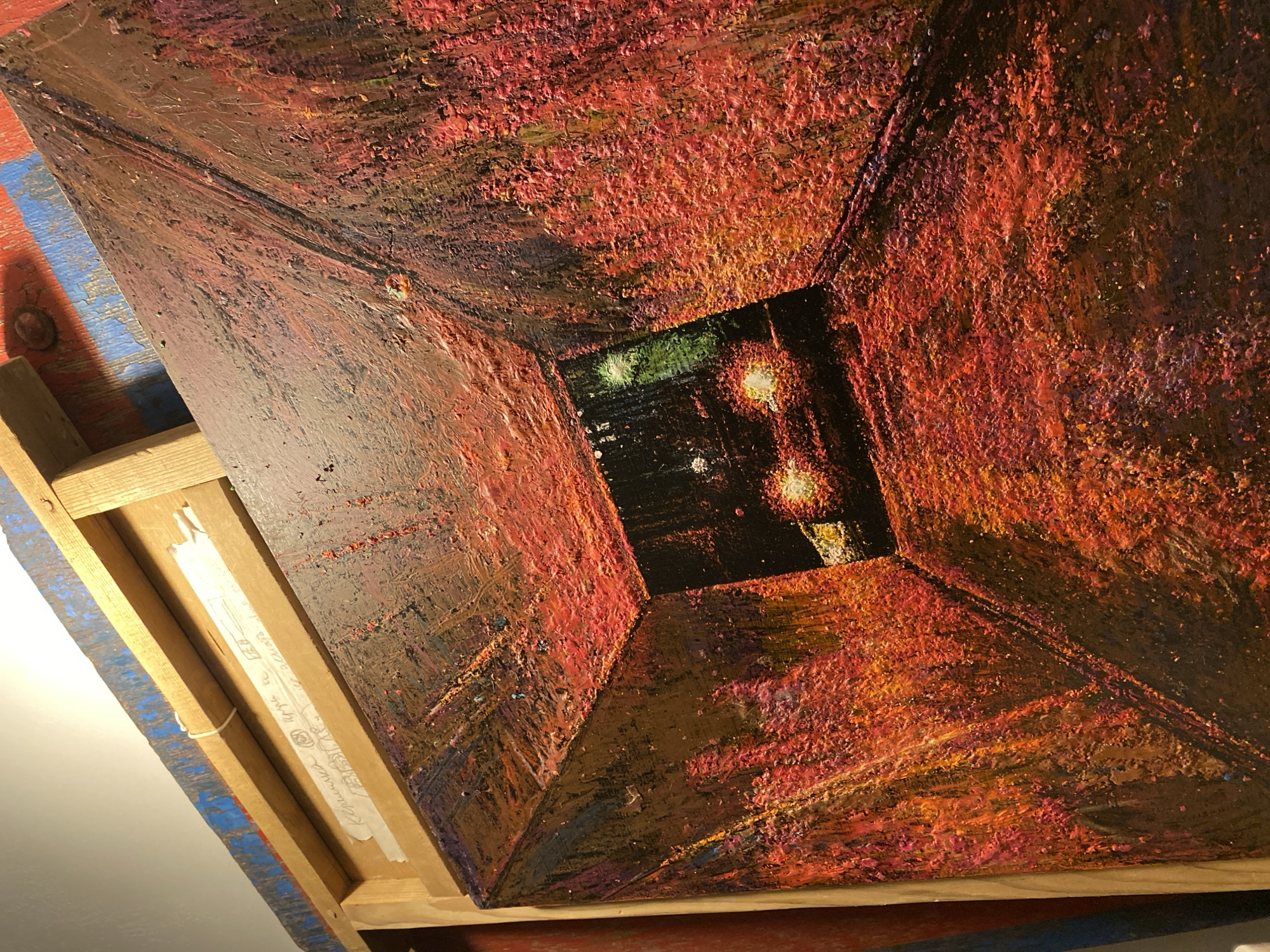 The image is a painting of a tunnel or underpass, possibly depicting a scene in Saint Petersburg, Russia. The artwork is rendered with a thick impasto technique, giving it a textured, almost sculptural quality. The dominant colors are fiery reds, oranges, and yellows, which create a sense of intense light and heat.
In the distance, at the end of the tunnel, are the headlights of what appears to be a vehicle. The lights are a bright yellow-white, cutting through the darkness. The walls of the tunnel are heavily textured with thick brushstrokes, creating a rough, uneven surface that catches and reflects the light. The perspective of the painting draws the viewer's eye into the deep vanishing point of the tunnel.
The frame of the painting is visible on the left side, made of unfinished wood. Tucked into the frame is a piece of paper with handwritten text in Cyrillic, likely a signature or inscription by the artist. The background suggests the painting is on an easel or leaning against a surface. The overall impression is a dramatic and emotive portrayal of an urban passage.