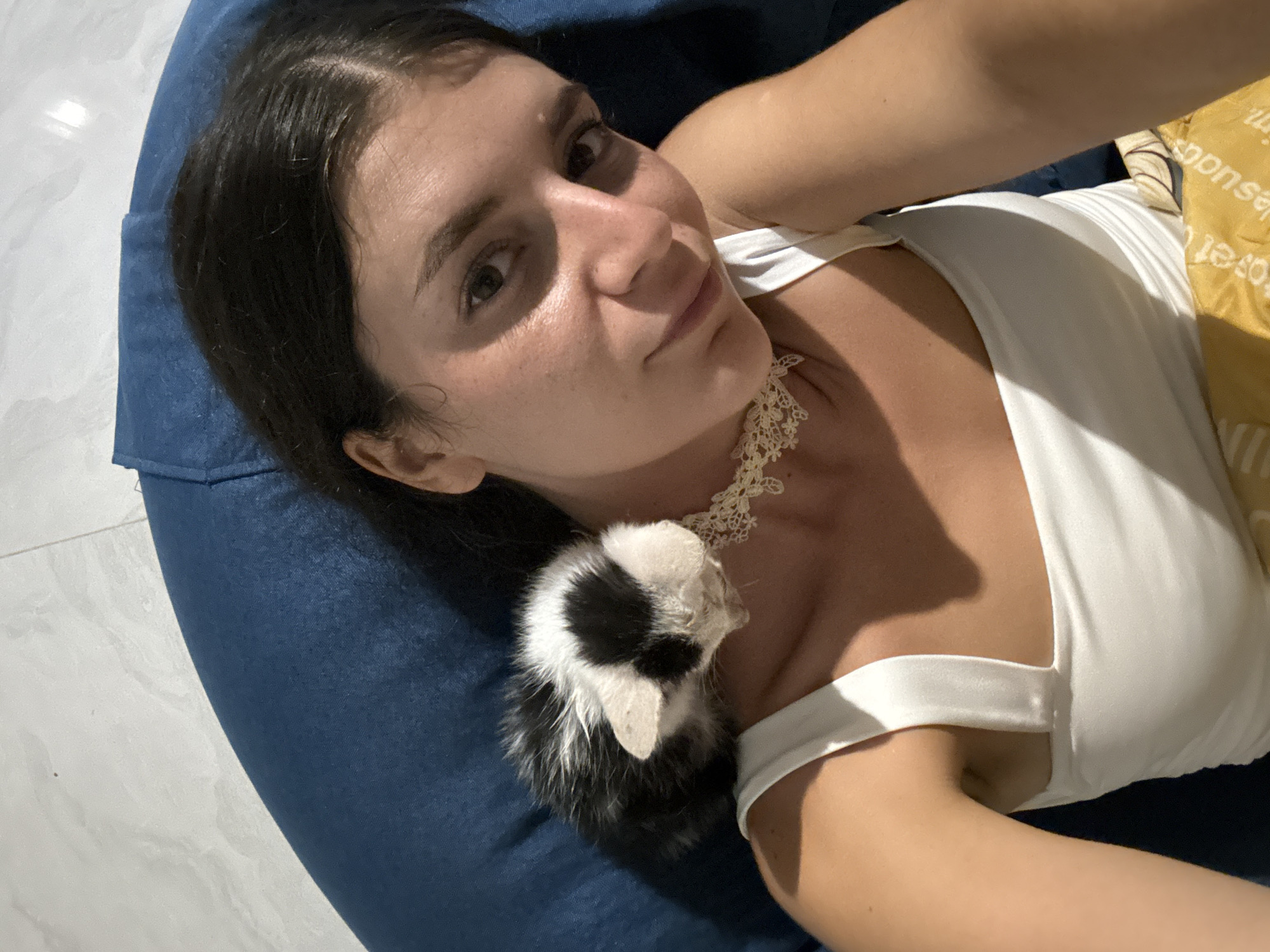An overhead shot shows a young woman with dark hair lying on a dark blue beanbag chair, with a small black and white kitten resting on her shoulder. The woman is wearing a white tank top and a delicate white lace choker necklace. Her face is turned slightly towards the camera, and she has a gentle smile. The kitten's head is lowered, appearing to be asleep. The background consists of the beanbag chair and a pale tiled floor. The scene takes place indoors, possibly in a guesthouse or hotel room in Mũi Né, Vietnam, indicated by the presence of a yellow patterned fabric, likely a beach towel, visible near the bottom of the frame.