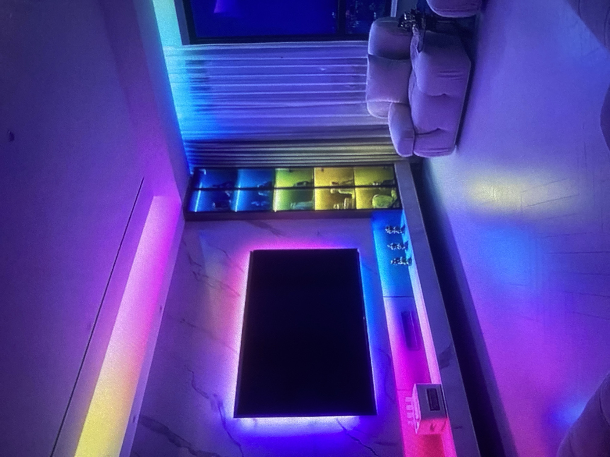 The image shows an interior space with colored LED lighting. A flatscreen television is mounted on a wall with marble-like paneling, illuminated by pink and blue LED lights. Above the television is a shelf with compartments, each lit with a different color: blue, yellow, and green. To the right of the television, there's a narrow shelf or display area with decorative objects and further LED lighting. A white, upholstered seating area is visible in the upper right corner of the frame, and a window with sheer curtains is above the shelf. The location is not possible to determine based on the image, so the city of Lhokseumawe, Indonesia cannot be confirmed.