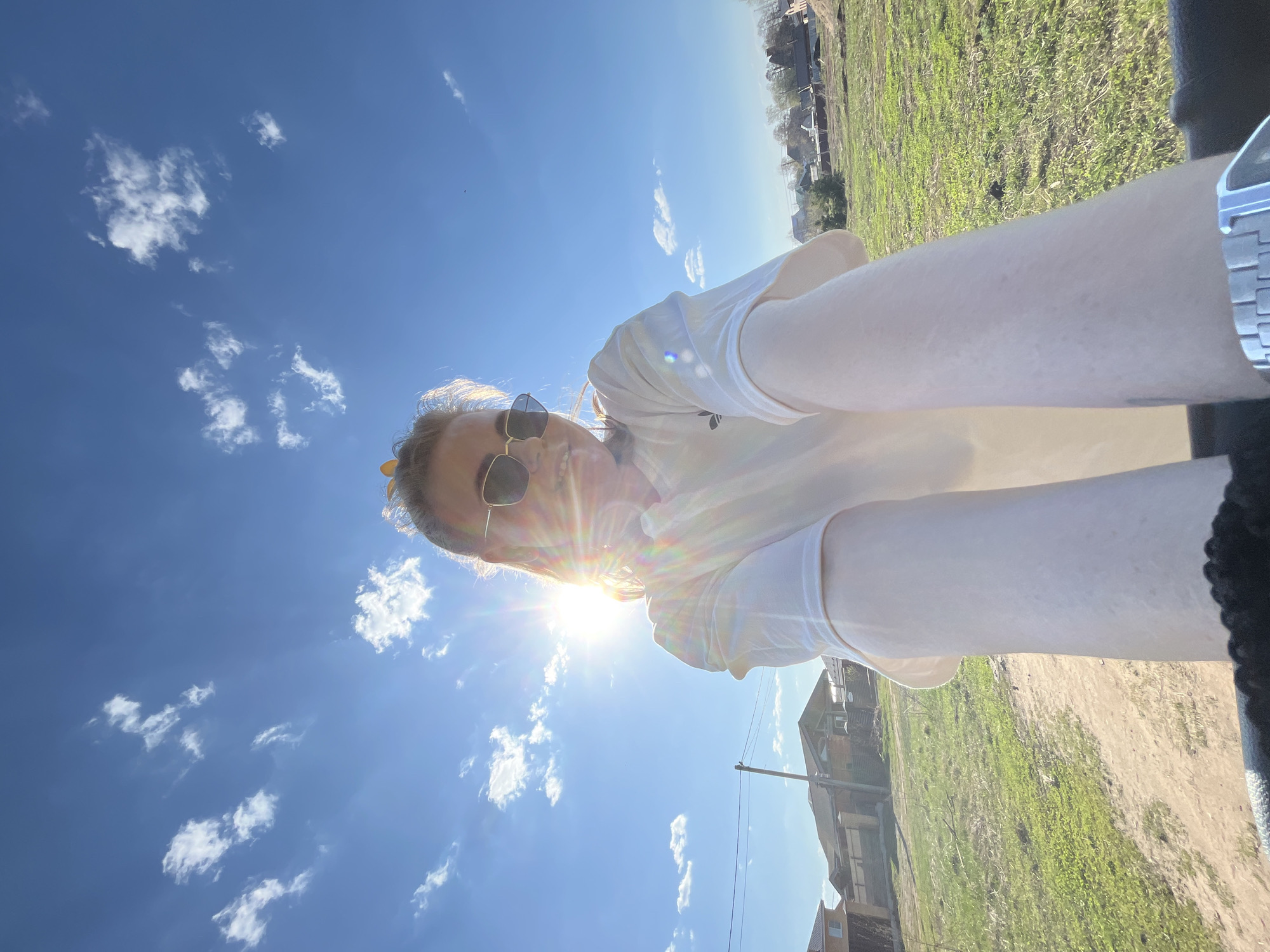 A low-angle, outdoor, daytime photograph shows a young woman with fair skin and light brown hair, wearing sunglasses and a white top, looking at the camera with a smile. The sun is directly behind her head, causing a lens flare. Above and to the left of her, there is a clear, blue sky with scattered white clouds. The background shows a grassy field, houses, and utility poles. Based on the architecture and vegetation, the image is likely taken in the city of Chaikovsky, Russia. The woman's left wrist is visible on the right side of the photo, where a silver watch can be seen. A dark object, possibly part of the car, is also visible next to the watch.