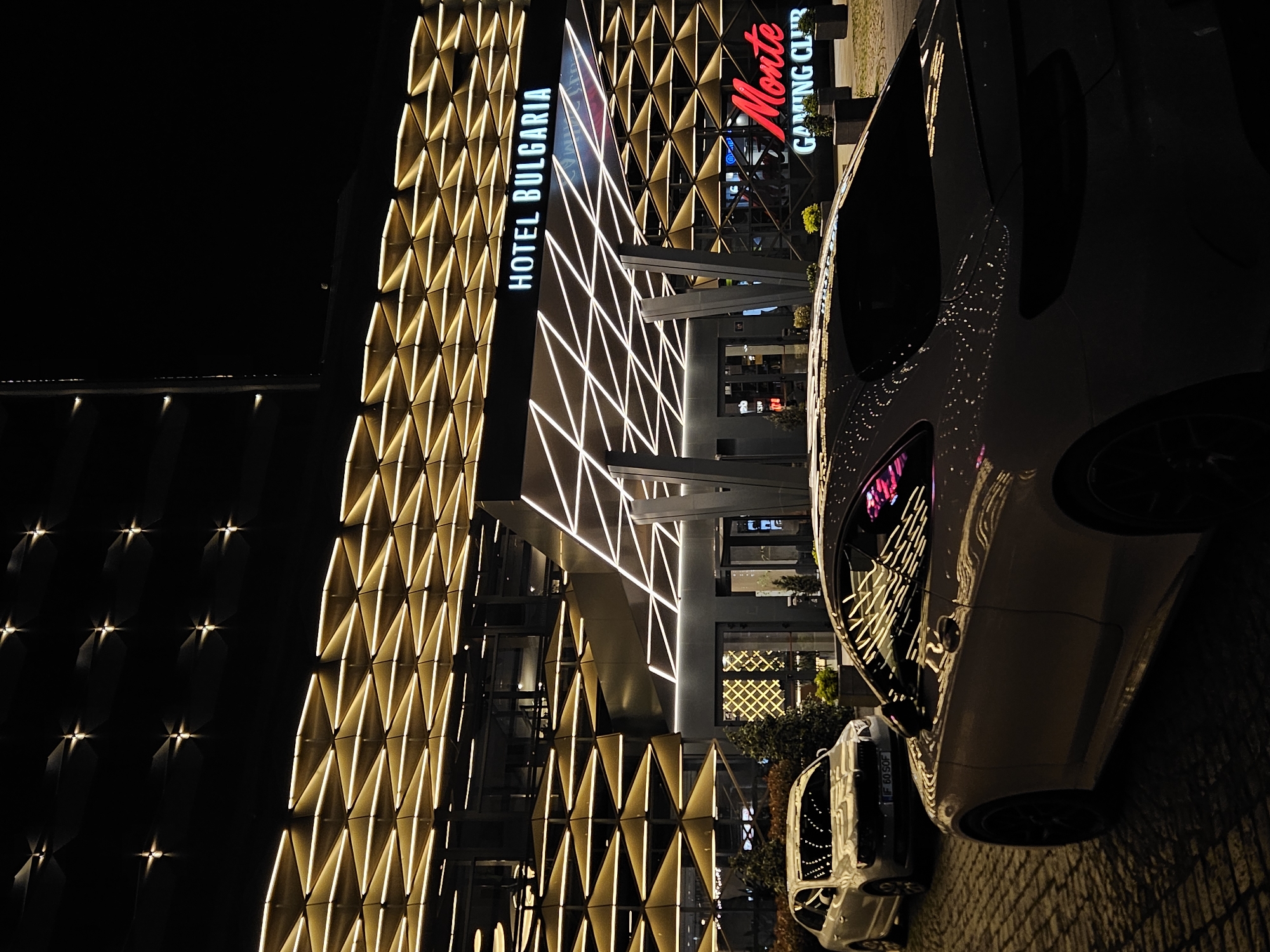 The image shows the Hotel Bulgaria and Monte Gaming Club in Burgas, Bulgaria at night. The hotel building is decorated with a geometric pattern of lights. A car is parked in front of the hotel. There are no people visible in the image.