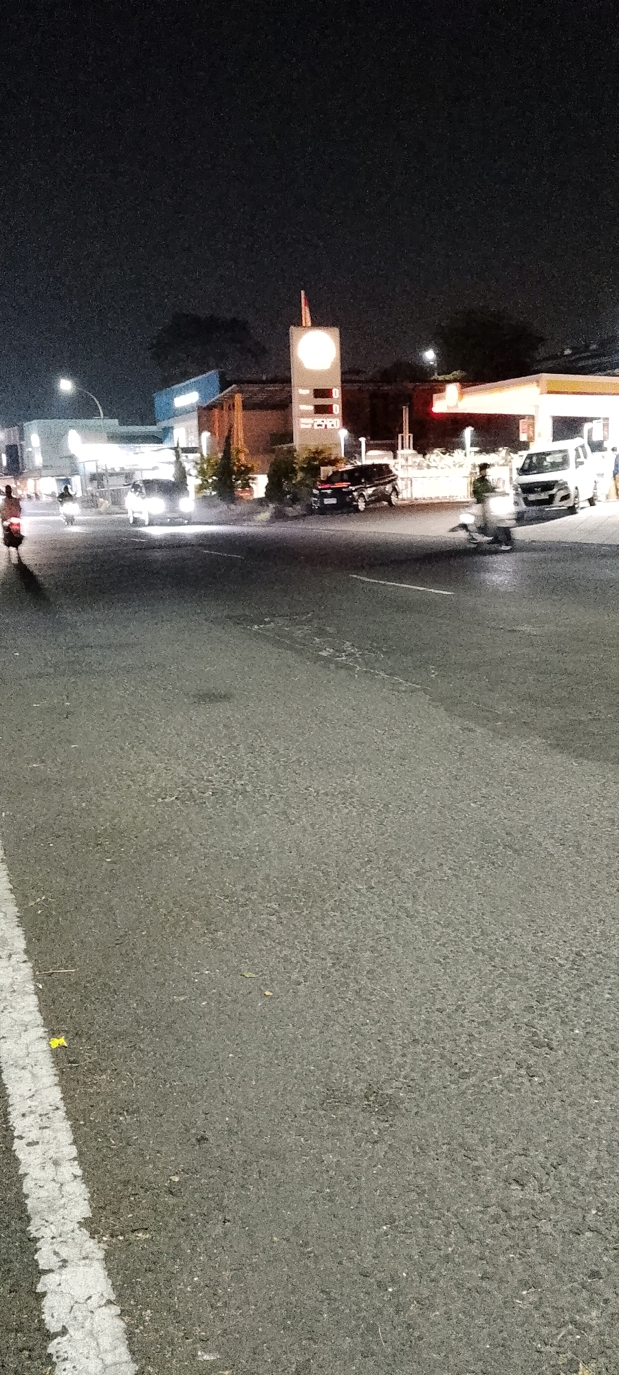 The image depicts a street scene at night in Cileungsi, Indonesia, likely near a gas station. A prominent Shell gas station sign with illuminated pricing is visible. Several vehicles, including a white van and a dark-colored SUV, are present on the road and at the gas station. A person on a motorcycle is seen in motion on the road, casting a shadow. Another individual is standing near the gas station pumps. The road is paved asphalt with a white line marking a lane. The background shows buildings and trees illuminated by artificial light, with a dark night sky above. The overall atmosphere suggests a typical commercial area operating at night.