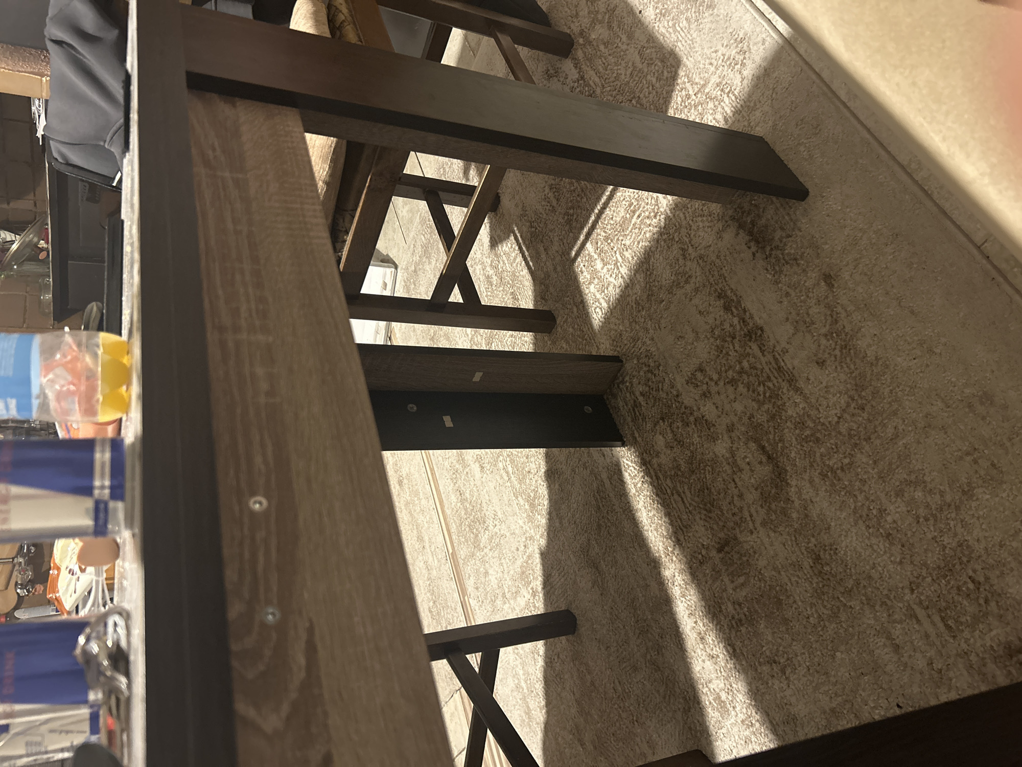 The image presents a low-angle, interior view beneath a wooden table in a domestic setting, located in Unknown, Unknown. The foreground shows the underside of a light-colored wooden table, displaying a visible grain pattern and secured by two prominent silver-colored fasteners. The dark, visible edge of the tabletop holds several objects: multiple tall, blue-labeled cans (identifiable as "DRINK" and "ENERGY D"), a clear plastic bottle containing yellow liquid, and a set of keys on a metallic keyring. A dark fabric item is draped over an object in the background on the tabletop.
Below the table, dark wooden chair legs and cross-bracing structures are visible, with a portion of a light-colored, upholstered chair cushion discernible on the right side. The floor is covered with a light beige, textured carpet, upon which strong, elongated shadows from the table and chair legs are cast, indicating a directional light source. A light-colored floor transition strip is visible along the bottom right edge of the frame. No individuals are present in the scene.