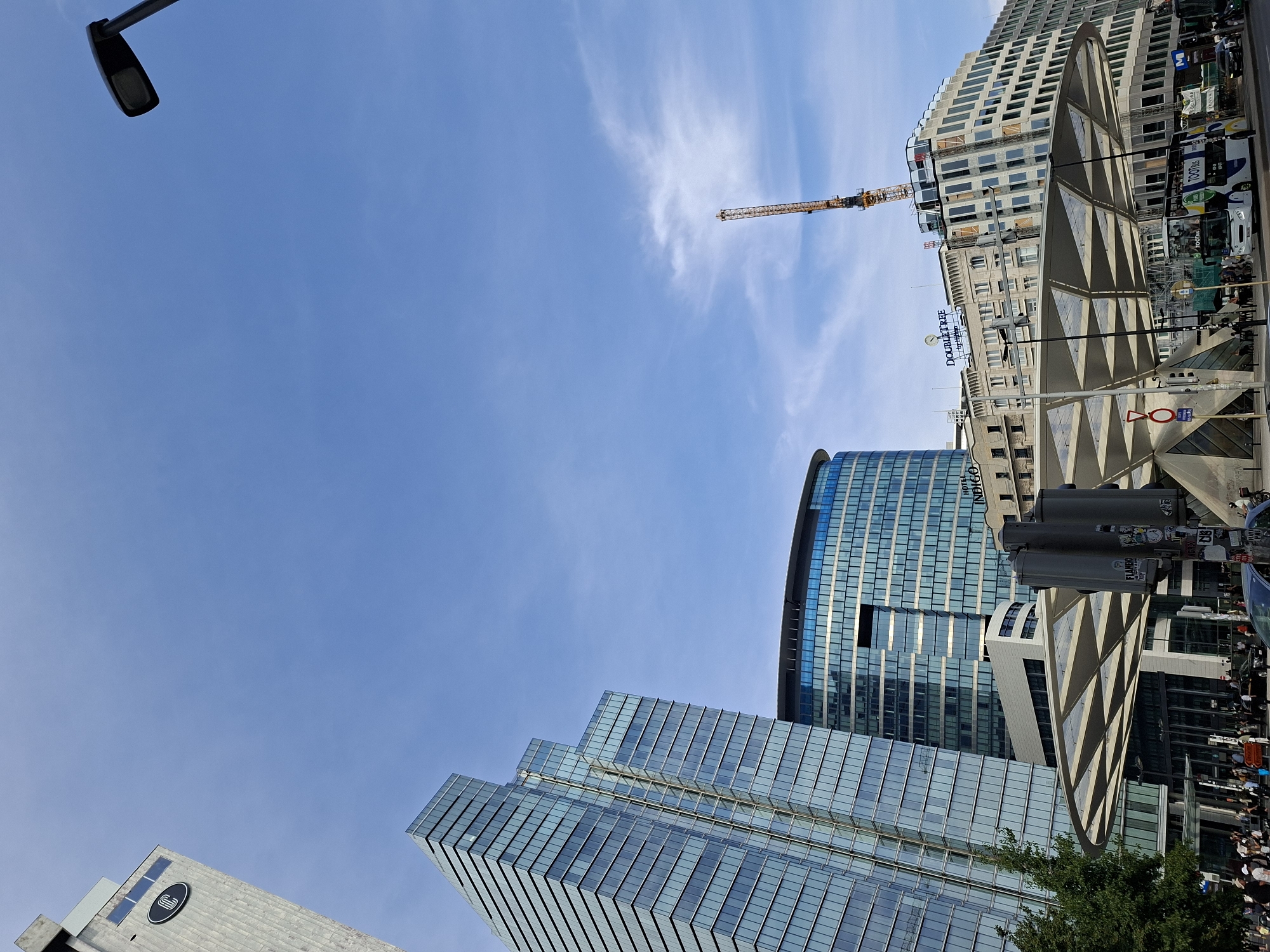 The image shows the skyline of Saint-Josse-ten-Noode - Sint-Joost-ten-Node, Belgium, dominated by modern glass skyscrapers and older architectural styles under a clear blue sky with wispy clouds. In the foreground, a futuristic-looking white canopy structure is visible, with a street and pedestrian activity below. Several people are walking and standing around the area. Traffic lights and street signs are present. A double-decker tourist bus is seen on the street. In the background, a construction crane is active, indicating ongoing development. The buildings include the Indigo Hotel and a Doubletree by Hilton hotel. One skyscraper has a circular logo with a "C" inside. The overall scene depicts a bustling urban environment with a mix of contemporary and historical architecture.