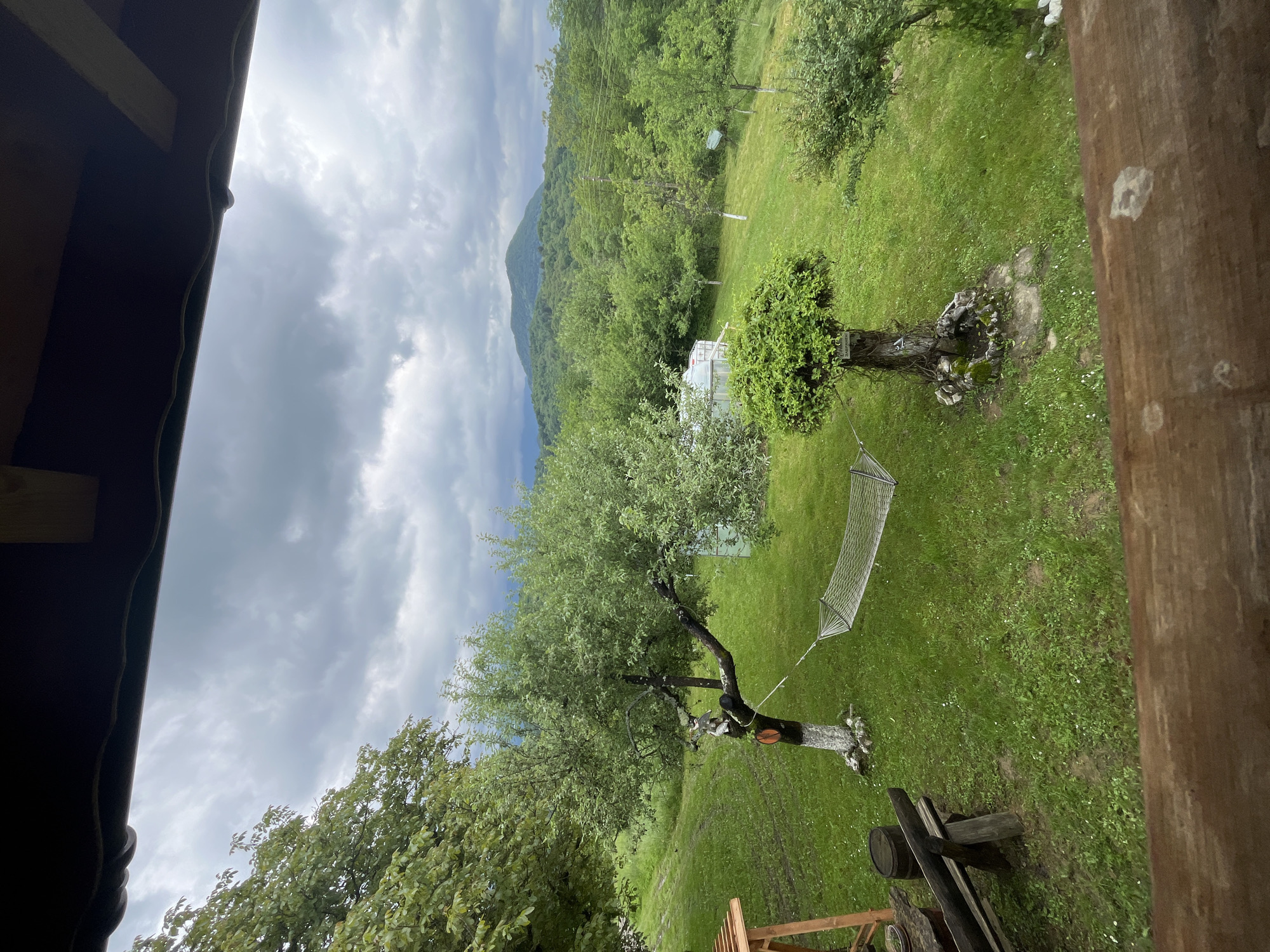 The image shows a view of a backyard or garden landscape in Shopite, Bulgaria. The scene is dominated by lush green grass and various trees, including a pruned tree near a hammock. A small structure, possibly a greenhouse or shed, is visible in the background. The sky is overcast with dark, stormy clouds. A mountain or hill is visible in the distance, covered with dense forest. The image appears to be taken from an elevated position, possibly a porch or balcony. Parts of the building structure, including the roof and wooden posts, are visible on the edges of the frame.