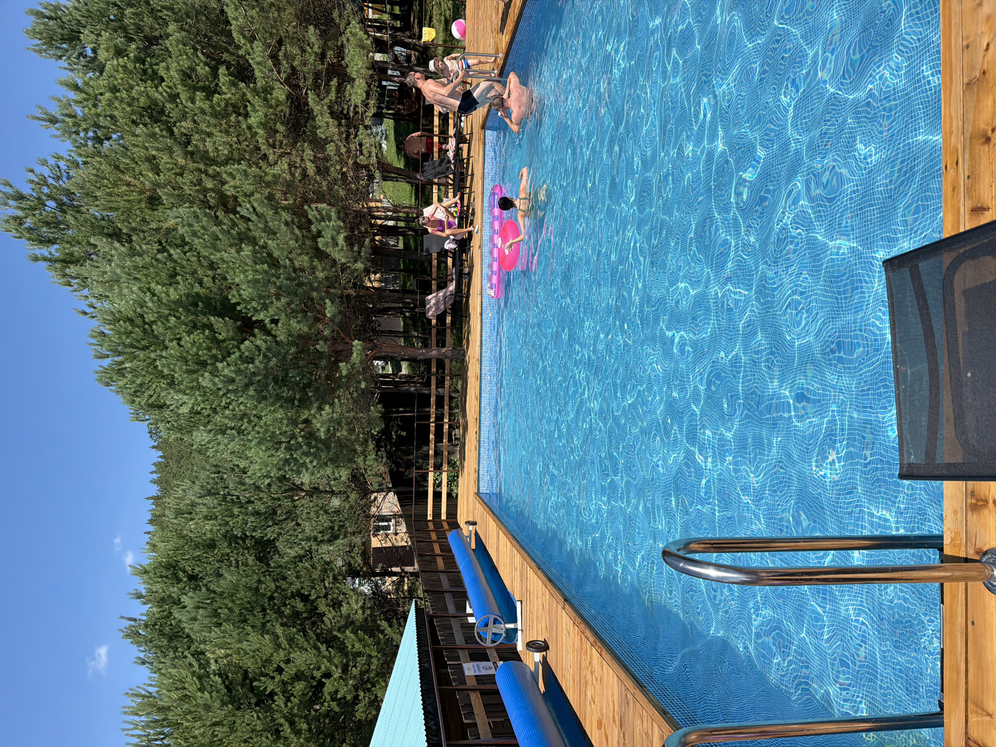 In Elekmonar, Russia, a crystal-clear blue swimming pool glistens under a bright, sunny sky. The pool, surrounded by a wooden deck, is partially occupied by several individuals enjoying the water. Three people are in the pool, two women and one man. One woman is lying on a pink inflatable flamingo, while another is in the water, facing away from the camera. A man is standing in the pool, holding onto the edge. Near the edge of the pool, a man in swim trunks is helping a young boy, who is wearing striped swim trunks, to get into the water. A pink inflatable ball rests on the deck nearby. In the background, lounge chairs are set up on the wooden deck, with some people seated and others standing. A wooden fence encloses the area, and behind it, a dense forest of tall pine trees stretches towards the horizon. The scene evokes a sense of summer leisure and relaxation at a resort or private residence in this scenic Russian location.