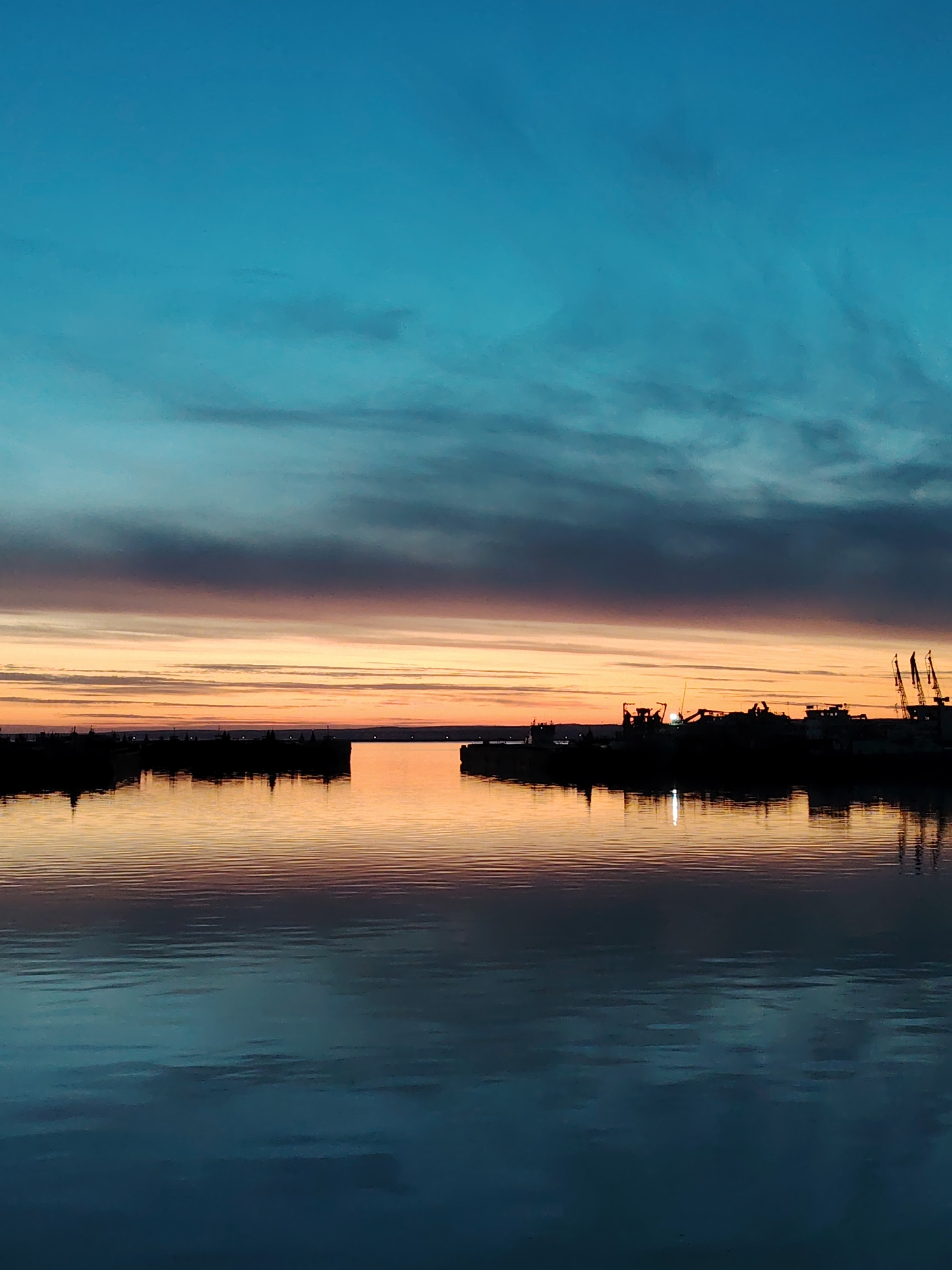 The image depicts a harbor scene at dusk. Silhouetted structures and cranes are visible on the horizon, their reflections mirrored on the still water. The sky transitions from a deep blue at the top to a gradient of orange and pink near the horizon. The lower half of the image is dominated by the water's surface, which exhibits subtle ripples and reflections. Based on visual cues, the location appears to be within the city of Chaikovsky, Russia, likely depicting the harbor area on the Kama River. No people are visible.
