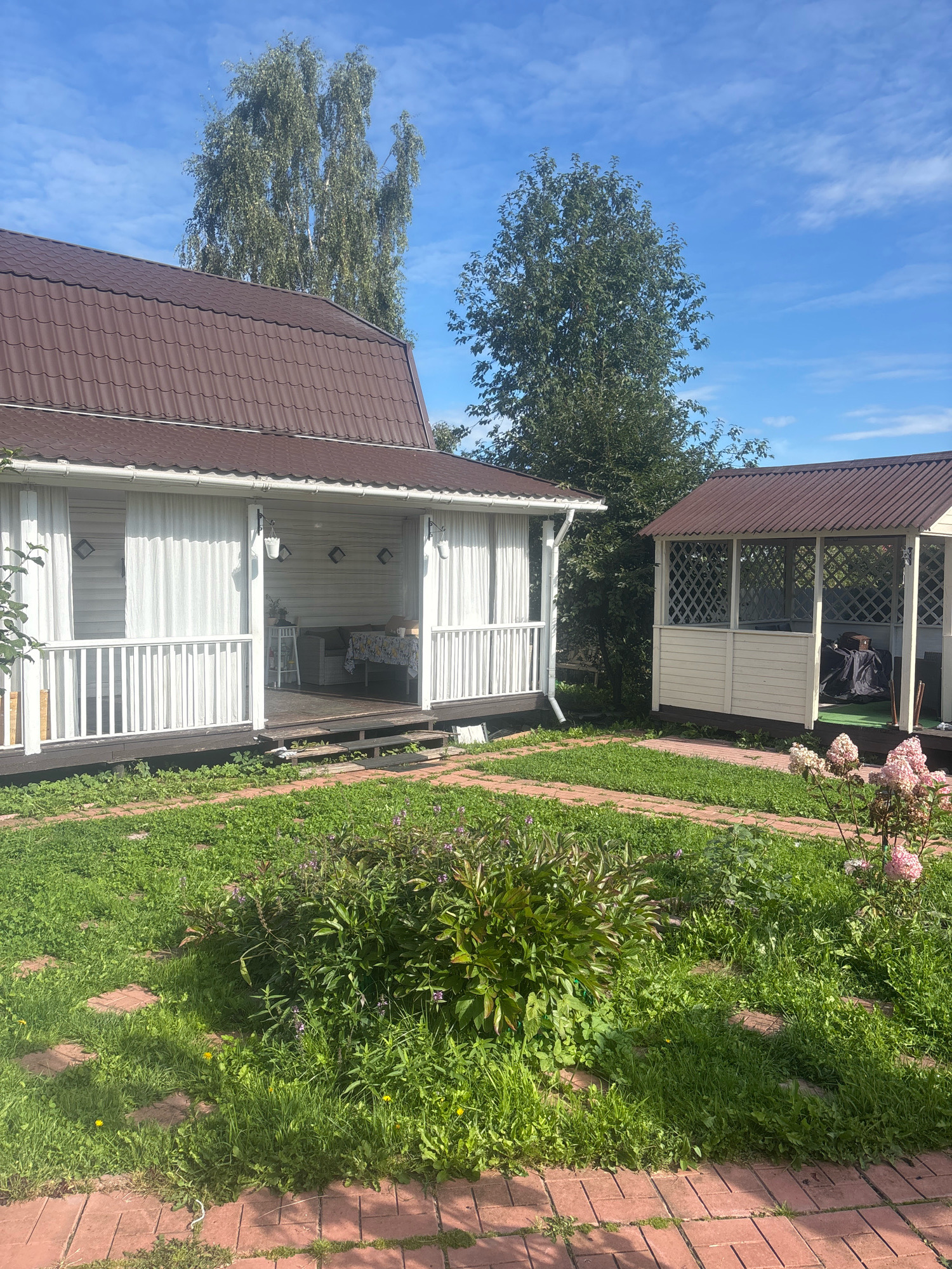 The image depicts a sunny day in the garden of a private residence in Лотос Снт (марьино), Russia. In the foreground, a red brick pathway winds through lush green grass and various plants, including a large green bush and a flowering pink bush. To the left, a white house with a brown metal roof features a covered porch with white railings and sheer white curtains. Furniture, including a table and chairs, is visible on the porch. To the right, a separate white gazebo with a brown metal roof stands in the yard. A large tree with green foliage is visible behind the house, and another smaller tree is partially obscured by the house. The sky is a bright blue with scattered white clouds. No people are present in the image.