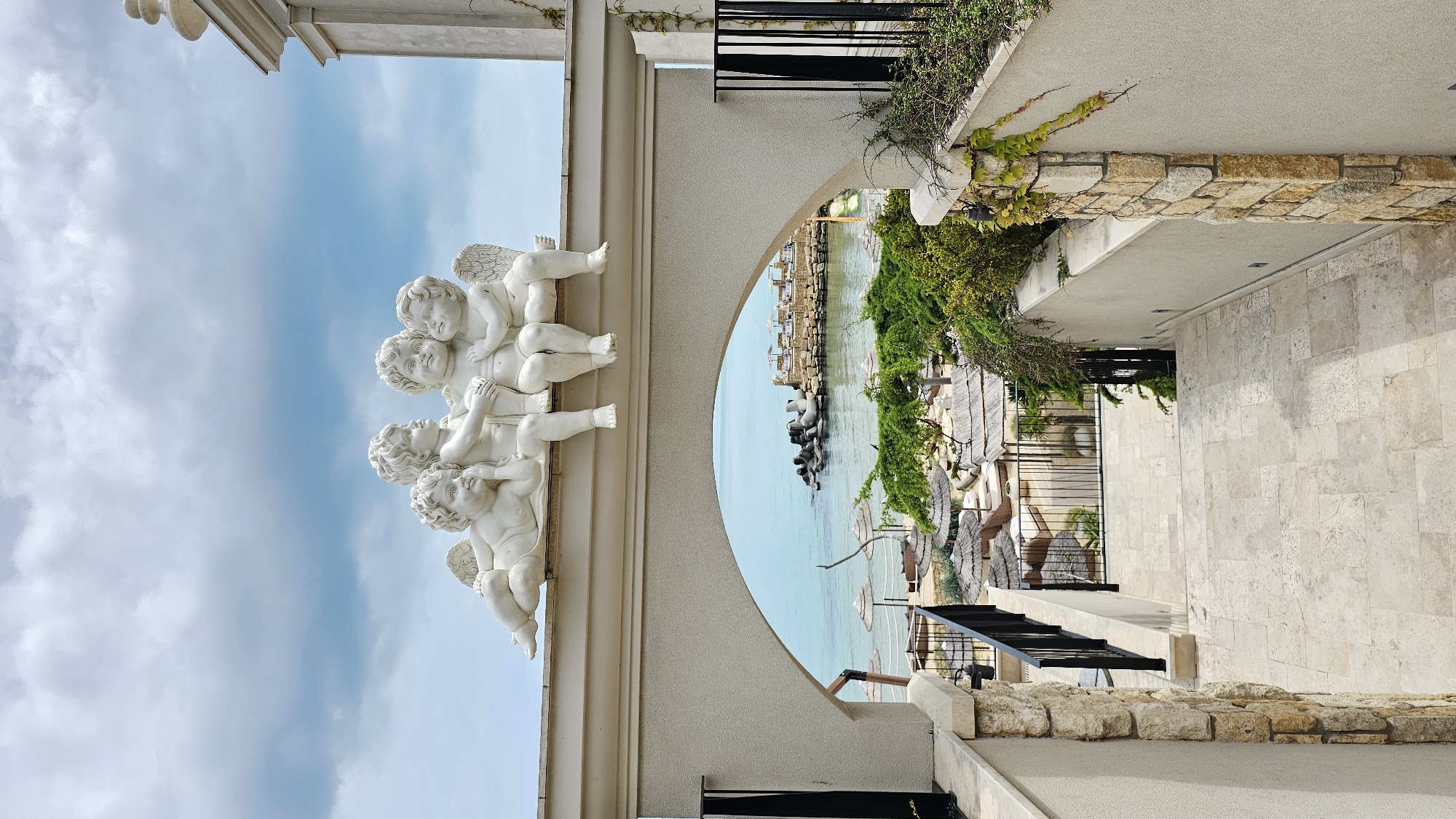 The image captures a view framed by architectural elements, likely in Varna, Bulgaria. On the left, a row of cherubic statues adorns a light-colored building facade, set against a blue sky with scattered clouds. Through an archway, a coastal scene is visible, featuring calm, turquoise water, beach umbrellas, and breakwater structures. The right side of the frame is occupied by a stone pathway and wall, with greenery cascading over the edge. The pathway appears to lead towards the coastal view. The scene is captured in daylight.