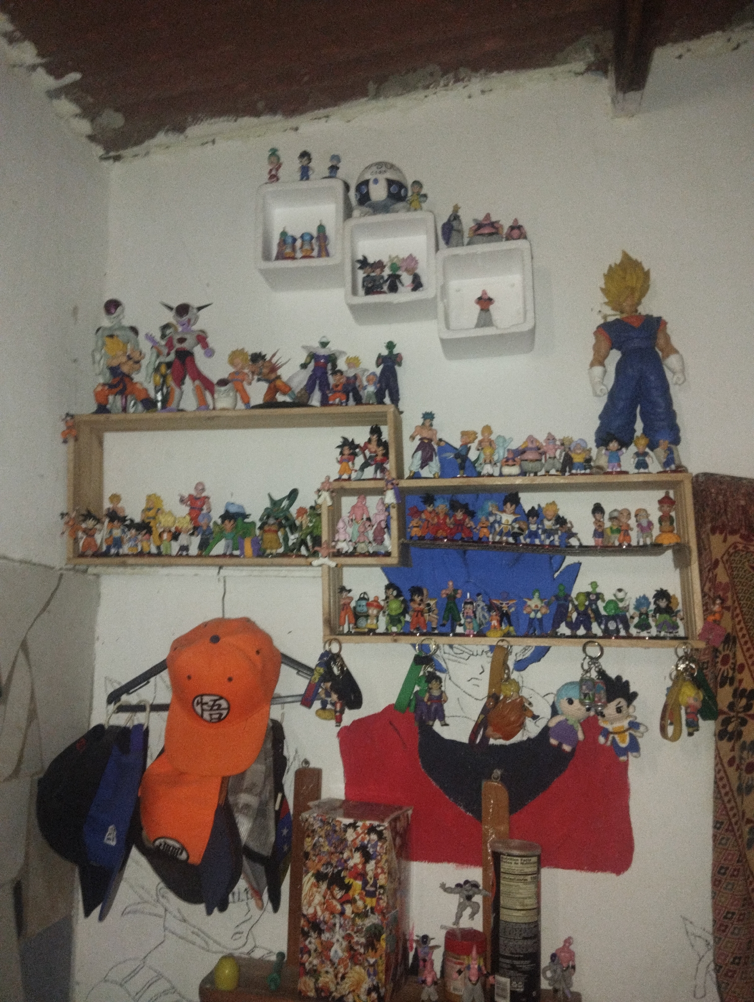 The image depicts a corner of a room, likely a bedroom, with a collection of Dragon Ball-themed items. The walls are white and show signs of wear. A wooden structure with shelves holds numerous small Dragon Ball figurines. Above the shelves, there are three white cube shelves also displaying figurines. An orange cap with a Dragon Ball symbol hangs on a coat rack, along with other caps. A red and black cloth is behind some of the items. Below, there is a table with a box filled with Dragon Ball images, figurines, and a nutritional supplement container. The room appears to be in a residential area, possibly El Valle del Espíritu Santo, Venezuela, based on geographic clues from the user. There are no people visible in the image, but the collection suggests a resident with a strong interest in Dragon Ball.