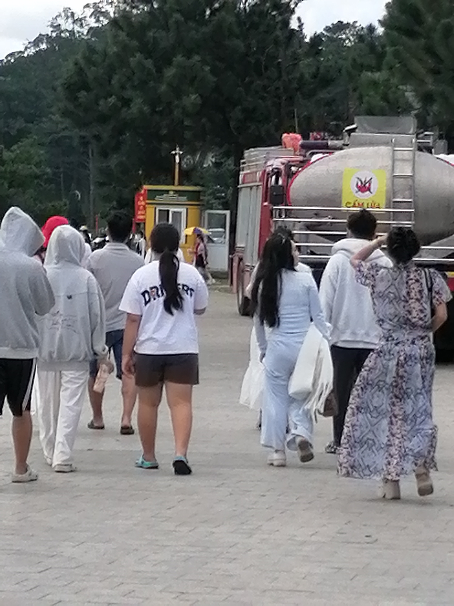 The image depicts a group of people walking outdoors on a paved pathway in Da Lat, Vietnam. In the background, a red and silver tanker truck with a ladder is visible, along with a small yellow building that appears to be a booth or kiosk. Trees form a dense backdrop. Several individuals are walking away from the camera. One woman in the foreground wears a white t-shirt with "DRIVER" printed on it and dark shorts. To her left, a person in a light blue tracksuit walks with a white bag over their shoulder. Further left, two individuals in hooded sweatshirts and white pants are walking. To the right of the woman in the "DRIVER" shirt, another person in a patterned dress is walking. The pathway appears to be made of interlocking grey pavers. The scene suggests a public gathering or event.