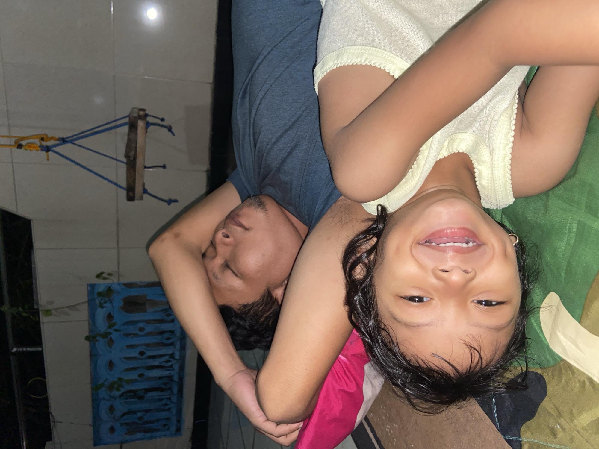 A father and daughter are lying together, presumably on a bed or soft surface. The image is oriented with their heads on the bottom and feet on the top. The father, on the left, has dark hair and is wearing a gray shirt. His eyes are closed, suggesting he may be sleeping or resting. The daughter, on the right, is smiling at the camera. She has dark hair and is wearing a light yellow shirt. Her arm is draped over her father's chest.
In the background, the tiled wall and clothesline apparatus give a clue that the location is in Gelam, Indonesia. A blue painted trellis is visible.