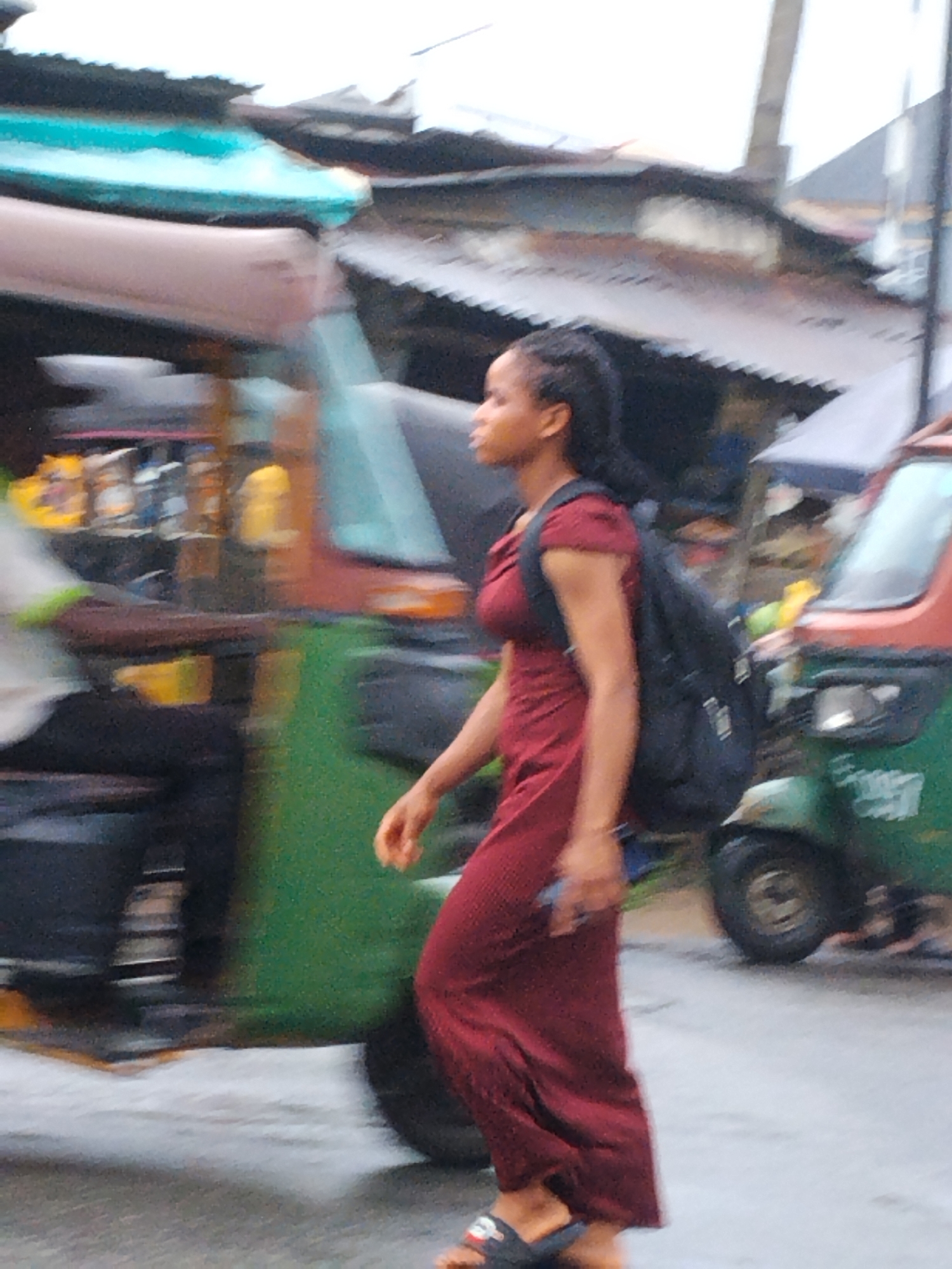 A woman with dark hair, a red dress, and a black backpack walks along a street in Aba, Nigeria. She is wearing sandals and is holding a phone in her left hand. Several auto rickshaws, locally known as kekes, are nearby. Buildings with corrugated metal roofs are visible in the background.
