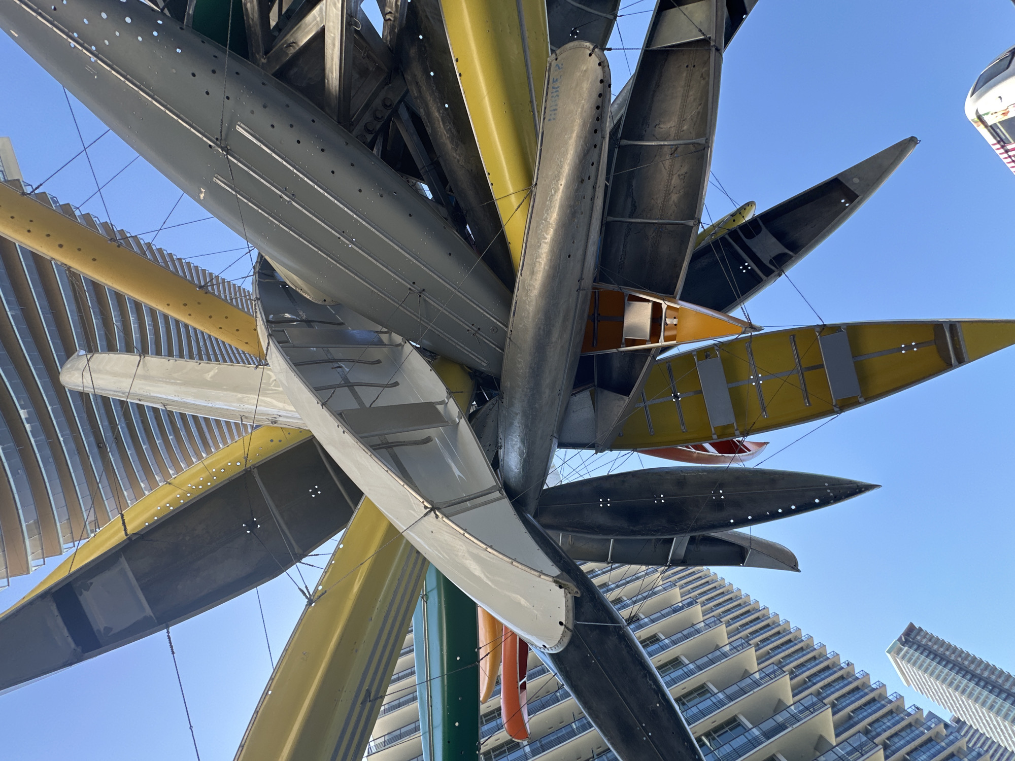 This is an upward-angle, full shot of an art installation composed of many canoes suspended in the air. The canoes are of various colors including yellow, gray, white, orange, green and black. They are arranged in a radial pattern. In the background, the curved facade of a modern building with rows of windows is visible, along with a clear blue sky. The building appears to be part of a cityscape. The location of the art installation is outside The Amazing Brentwood mall in Burnaby, Canada. There are no people visible in the image.