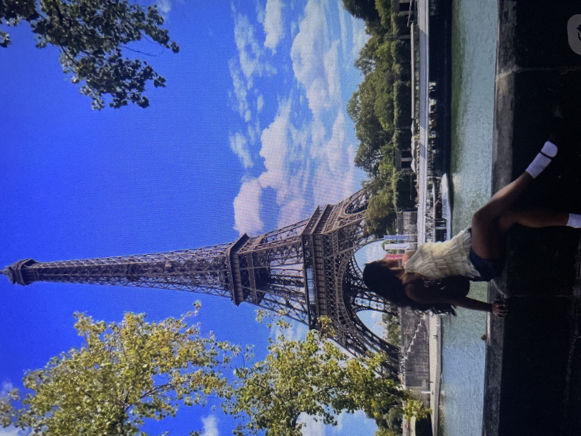 This is a full-frame, eye-level, outdoor, daytime photograph of the Eiffel Tower in Paris, France. The Eiffel Tower is centrally located in the background, dominating the scene. A person with long, dark hair is sitting on a stone wall in the foreground, facing to the right and looking towards the Eiffel Tower. The person is wearing a light-colored, textured top and denim shorts, with white socks and dark shoes. A dark bag is slung over their shoulder. A body of water, likely the Seine River, is visible in the middle ground, with a boat passing by. Trees and greenery are present on the far bank of the river and also frame the upper left and right corners of the image. The sky is a clear, bright blue with scattered white clouds. There is no indication of the city of Kołobrzeg, Poland in this image.