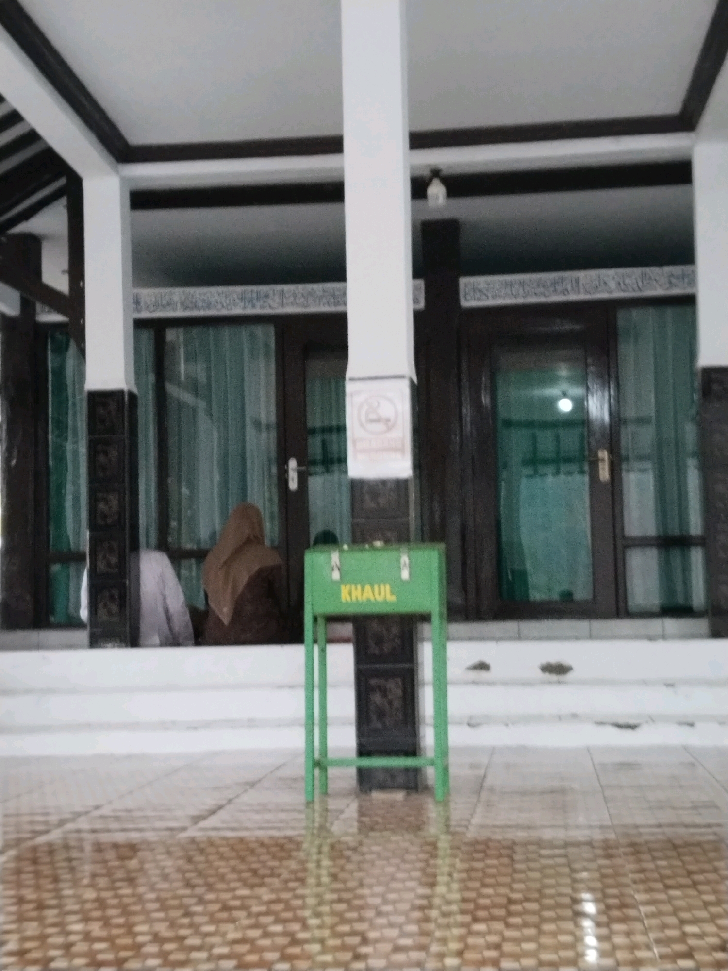The image captures the exterior of a building, possibly a mosque, located in Sangkanjaya, Indonesia. The architecture features white pillars, dark brown trim, and a white ceiling. Through open doorways and windows, green curtains are visible. Inside, two individuals are seated, facing away from the camera; one wears a white garment, and the other a brown headscarf. In the foreground, a green donation box with the word "KHAUL" written on it is prominently placed. The ground is tiled with a pattern of brown squares. The overall scene suggests a place of worship or community gathering.