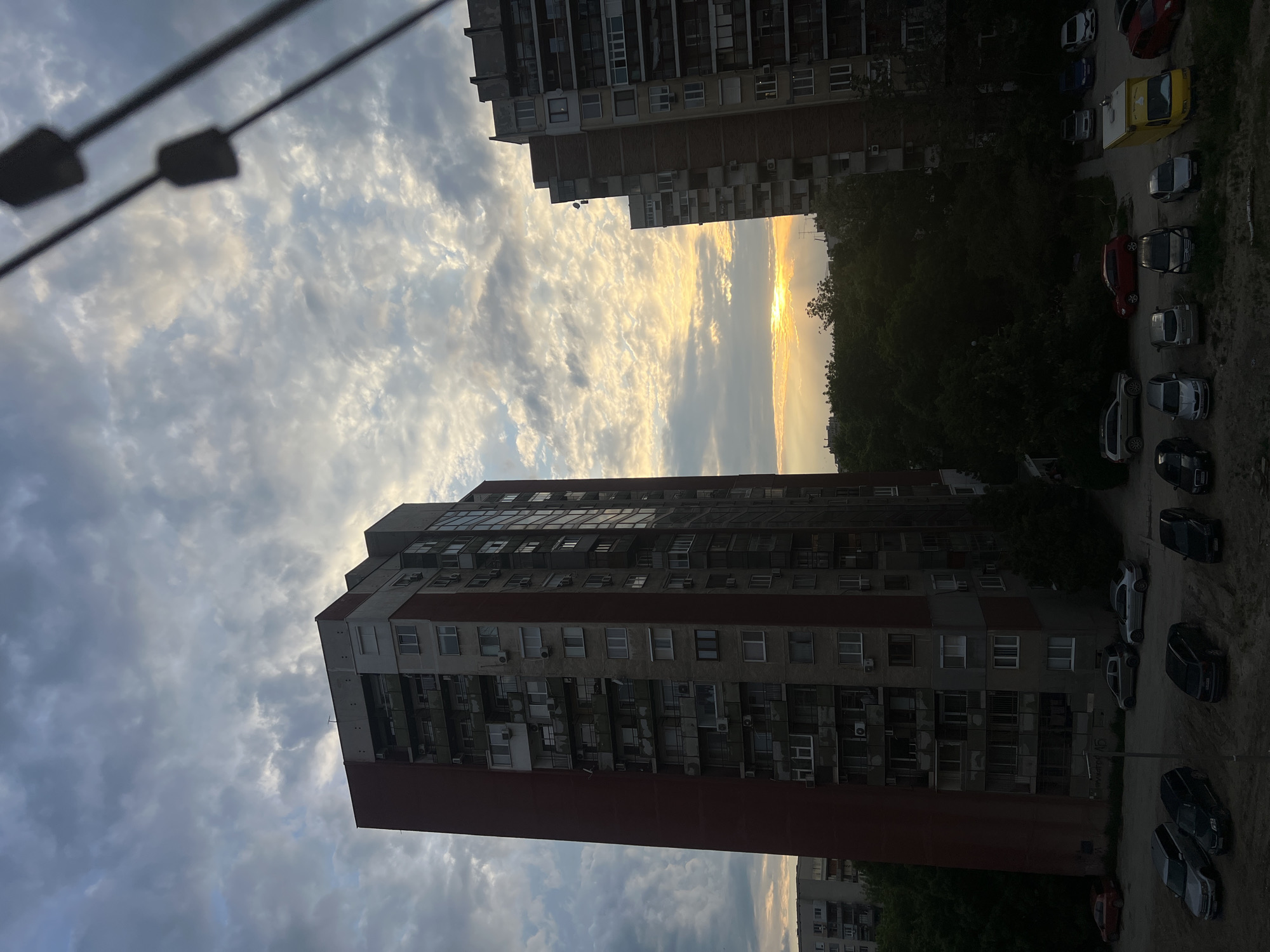A slightly rotated, wide-angle shot presents a view from an elevated position, likely a high-rise building. The perspective looks down at two tall, apartment buildings flanking a patch of trees, through which a golden sunset shines. To the right, a row of parked cars lines the side of one building. Overhead, wispy, textured clouds fill the sky. A streetlamp juts into the frame at the top left. The location is Unknown, Unknown.