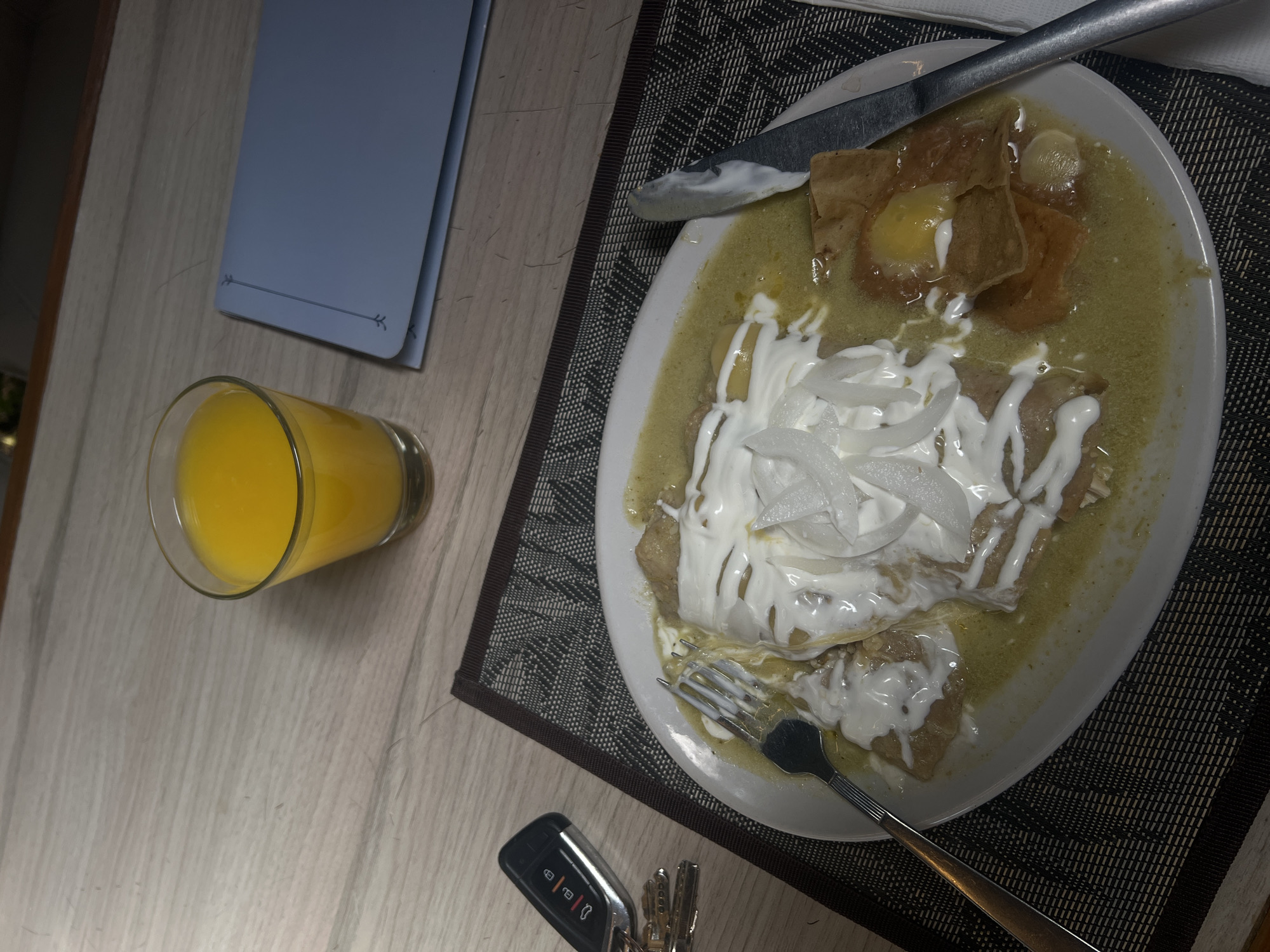 The image shows a plate of enchiladas with green sauce, cream, and onions, accompanied by a glass of orange juice, a fork, a knife, and car keys on a table with a placemat and notebook. The location cannot be determined from the visual information alone.