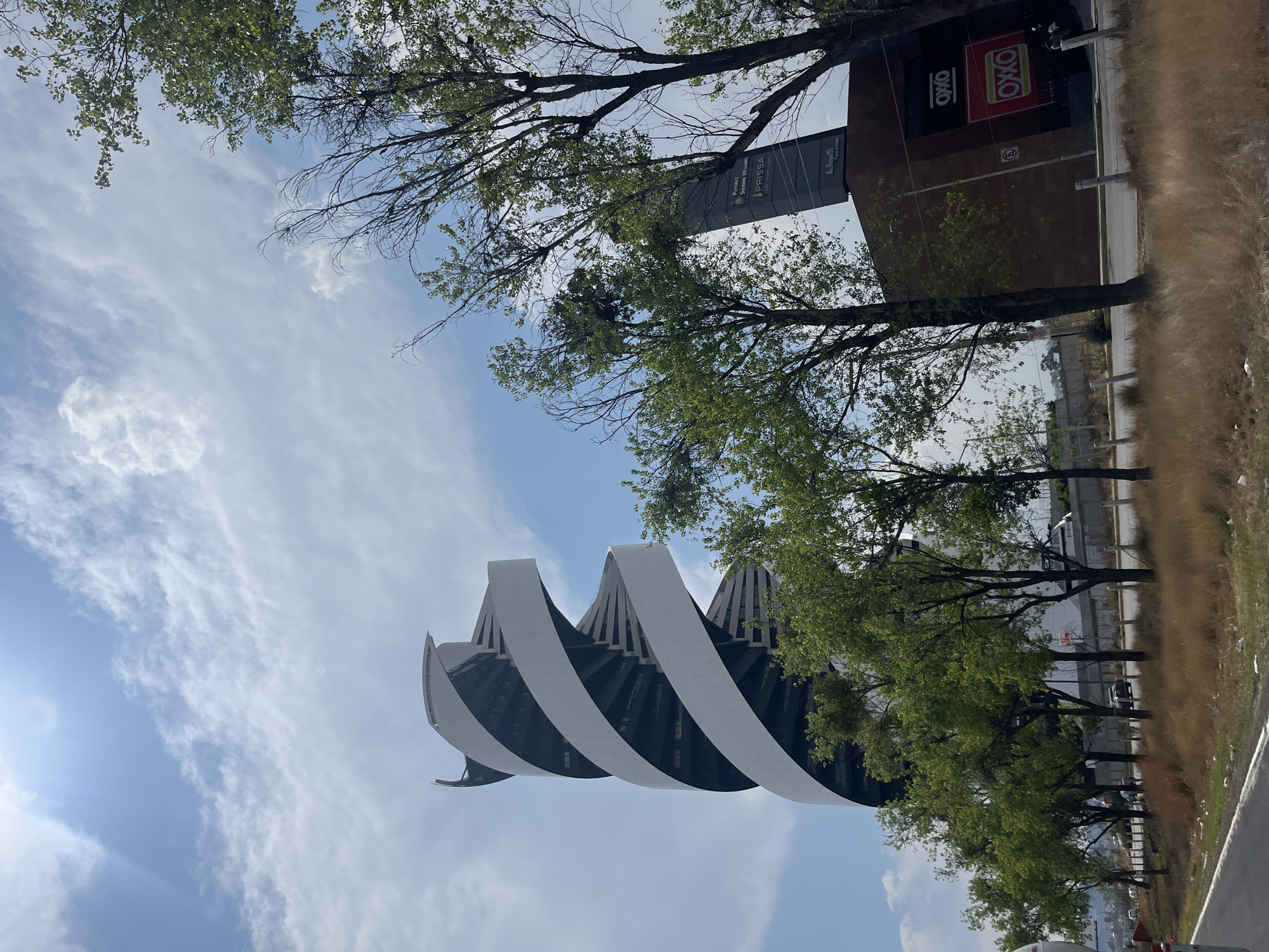 The image depicts the Museo Internacional Barroco (International Baroque Museum) in San Bernardino Tlaxcalancingo, Mexico, viewed from a low angle, emphasizing its distinctive architectural design. The museum's structure features a series of curved, white concrete forms spiraling upwards, contrasted against a blue sky with scattered clouds. Trees with green foliage partially frame the building, adding depth to the composition. Adjacent to the museum are commercial buildings, including an OXXO convenience store and a sign for PRISSA. A roadside with foliage and a glimpse of a road and a wall are also visible. There are no people visible in the image.