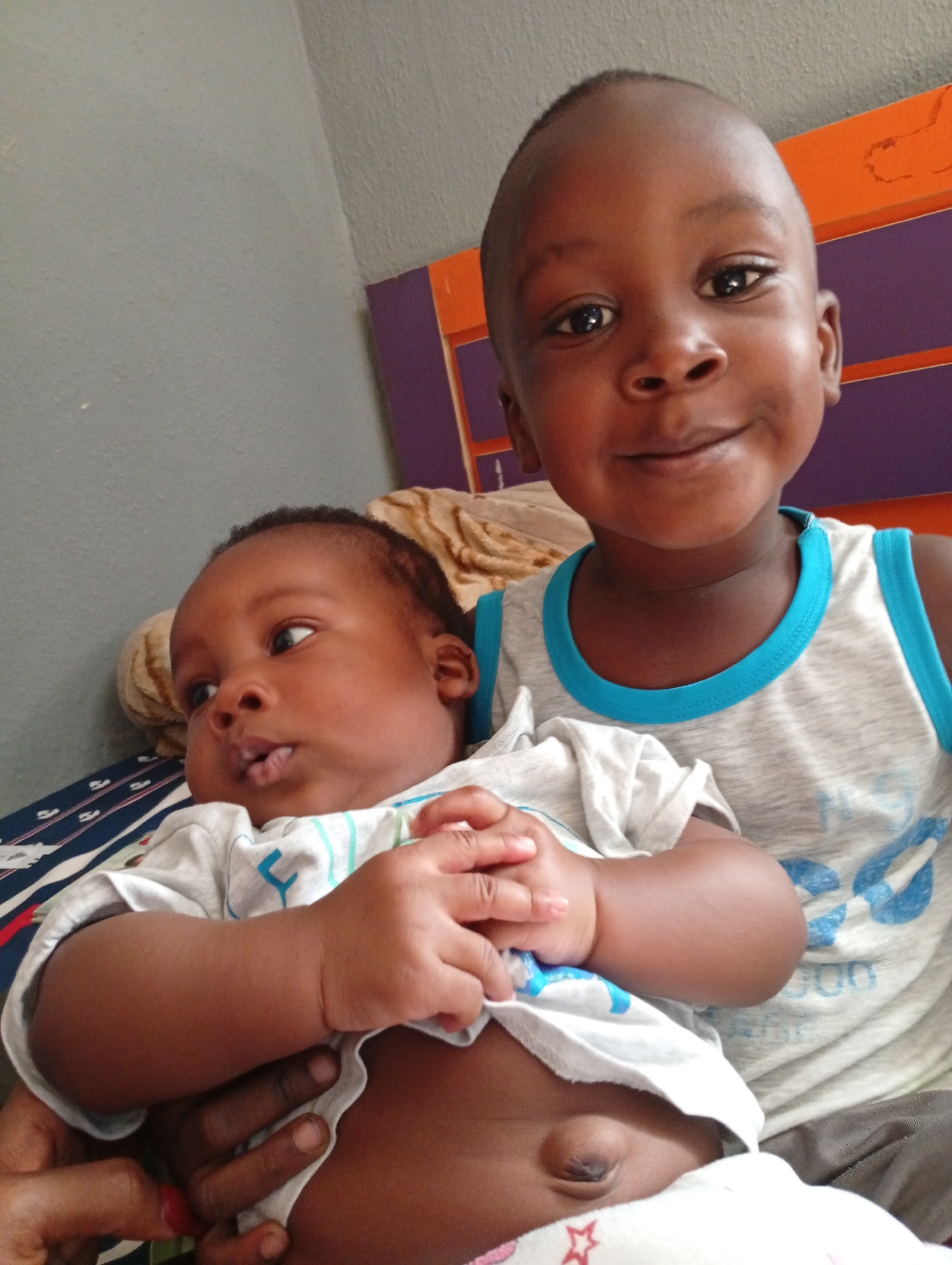The image shows two young, dark-skinned children. The older child, possibly a toddler, is standing behind and slightly to the right of the younger child, who appears to be an infant. The infant is being held in someone's arms and is wearing a light-colored shirt that is slightly pulled up, exposing part of the abdomen. The older child is wearing a gray tank top with a light blue trim around the neck and armholes.
The background includes a gray wall and a headboard with orange accents. A patterned blanket or sheet is visible.
The location is inferred to be Sabon Tasha, Nigeria, based on the provided context, but this is not definitively apparent from the image itself.
