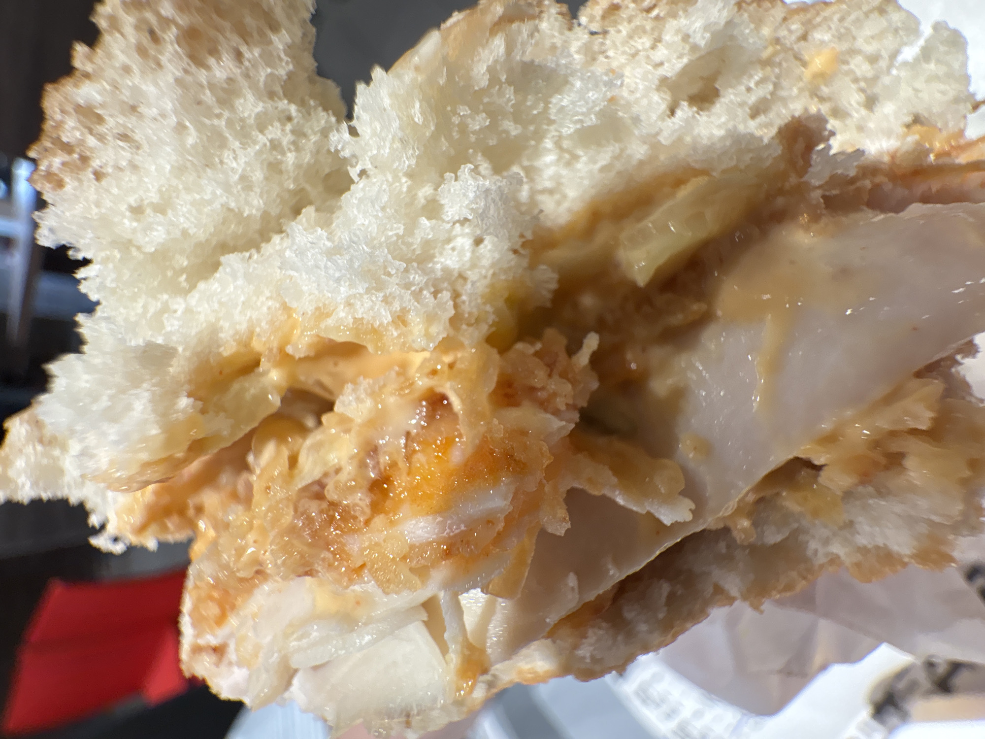 The image shows a close-up of a partially consumed sandwich. The sandwich consists of white, porous bread, possibly a roll or bun. Inside the bread, there are layers of sliced poultry (likely chicken or turkey), a yellowish-orange sauce or spread, and possibly some chopped pickles. The sandwich appears to be held in someone's hand and is resting on a white paper wrapper. The background is blurry but shows a red object (possibly part of a car or other object) and a metal railing or fence. There is no way to determine the location or connection to the city of Izhevsk, Russia from the image.