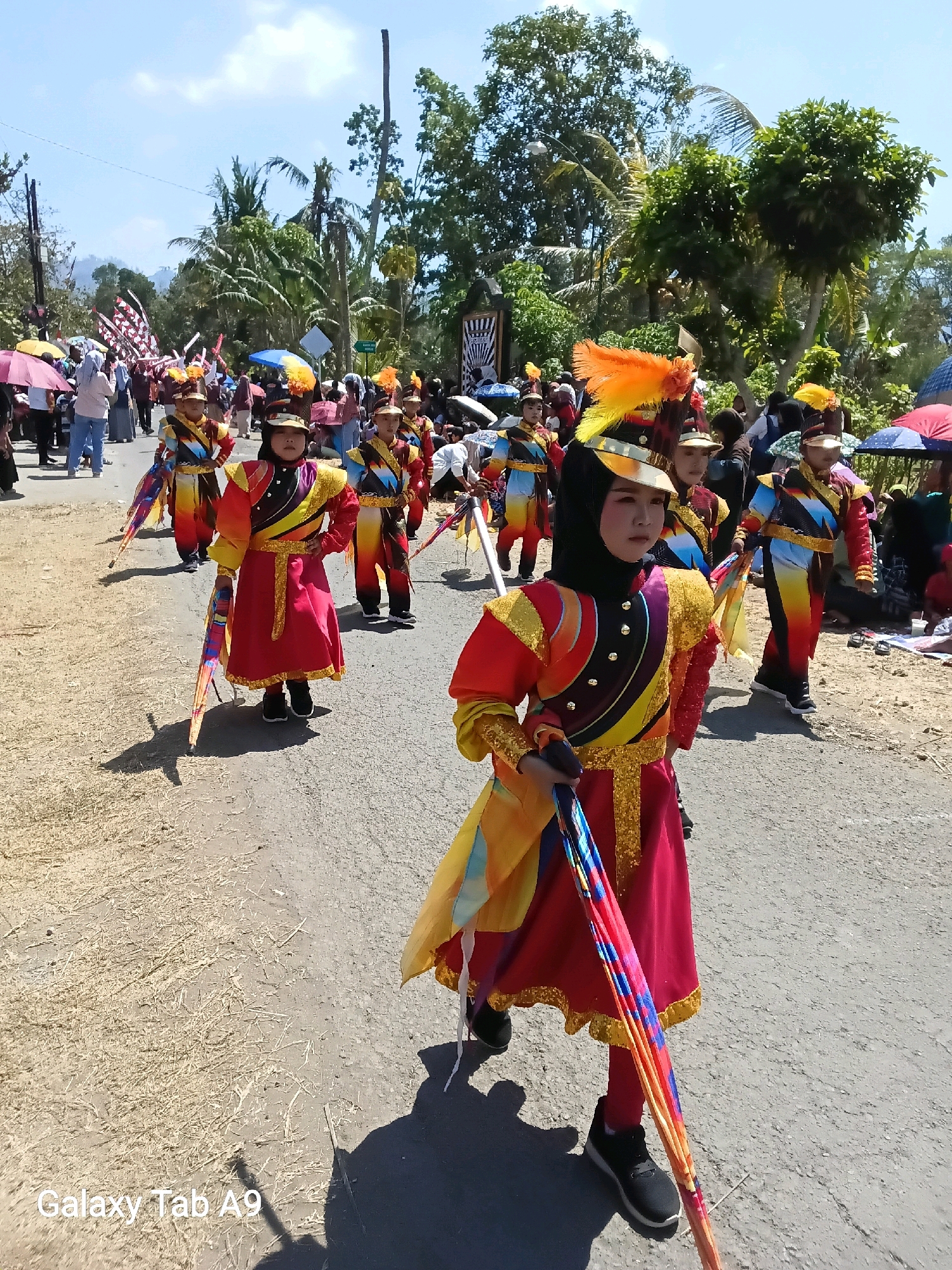 A group of children in colorful marching band uniforms are participating in a parade on a paved road in Ngadirenggo, Indonesia. The children are wearing red, yellow, and blue outfits with gold trim, and some have feathered hats. They are carrying decorative staffs or poles, and the child in the foreground is holding a folded umbrella with a vibrant pattern. Spectators are lined up along the sides of the road, some holding umbrellas for shade. Palm trees and other lush greenery are visible in the background, suggesting a tropical or subtropical environment. The sky is clear and blue, indicating a sunny day. The overall atmosphere appears festive and celebratory.