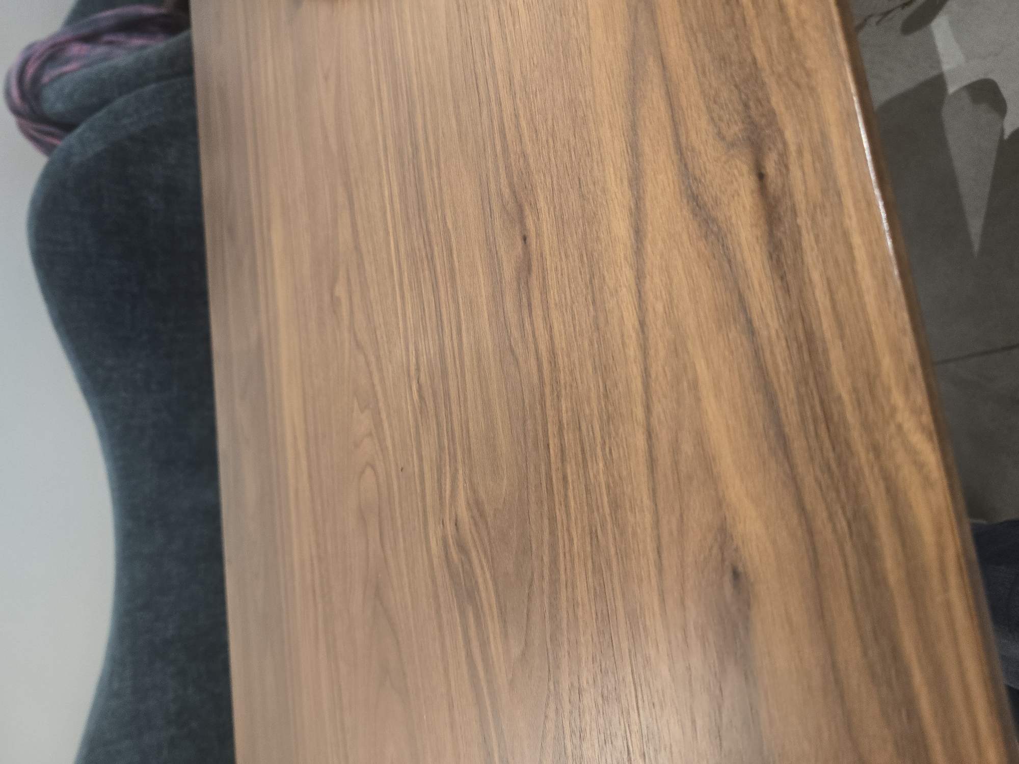 The image shows a close-up view of a wooden tabletop. The wood grain is prominent, with a warm brown color and natural variations. In the background, a dark blue upholstered furniture item is visible, with some purple and pink yarn draped over it. The setting appears to be indoors, possibly a living room or lounge area. There is no indication of the city of Ulan-Ude, Russia.
