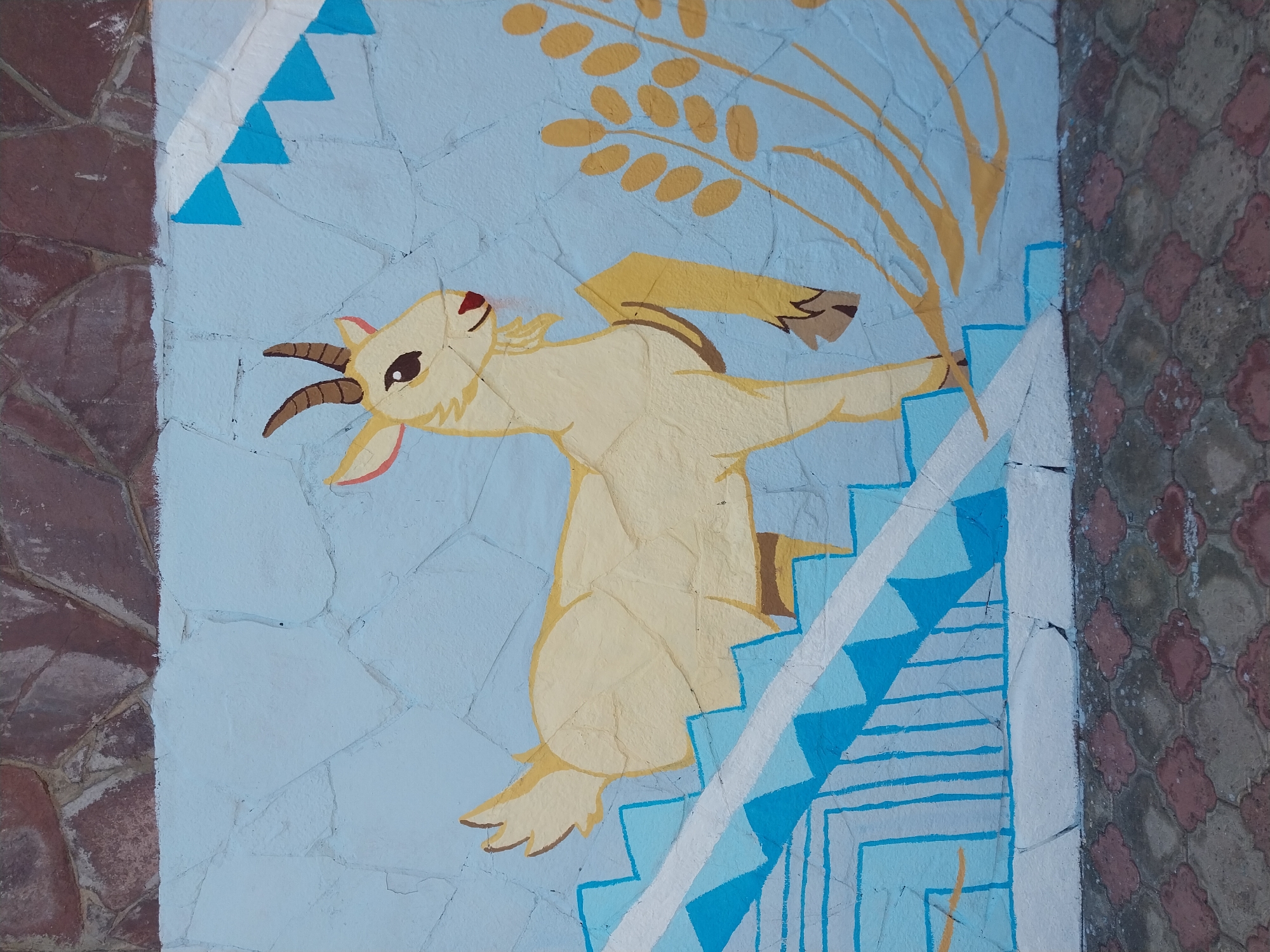 The image shows a mural painted on a light blue, textured wall, likely located in Orenburg, Russia. The mural depicts a light yellow goat with brown horns and hooves, standing on a blue and white staircase. The goat is facing right, with its tail raised. To the right of the goat, there are yellow stalks of wheat. Above the goat, there are blue triangular shapes and a white stripe. The wall itself is cracked and appears to be made of large stones. Below the wall is a paved area with red and gray interlocking pavers. The overall scene is outdoors, with the top of the image showing a darker stone wall above the mural.