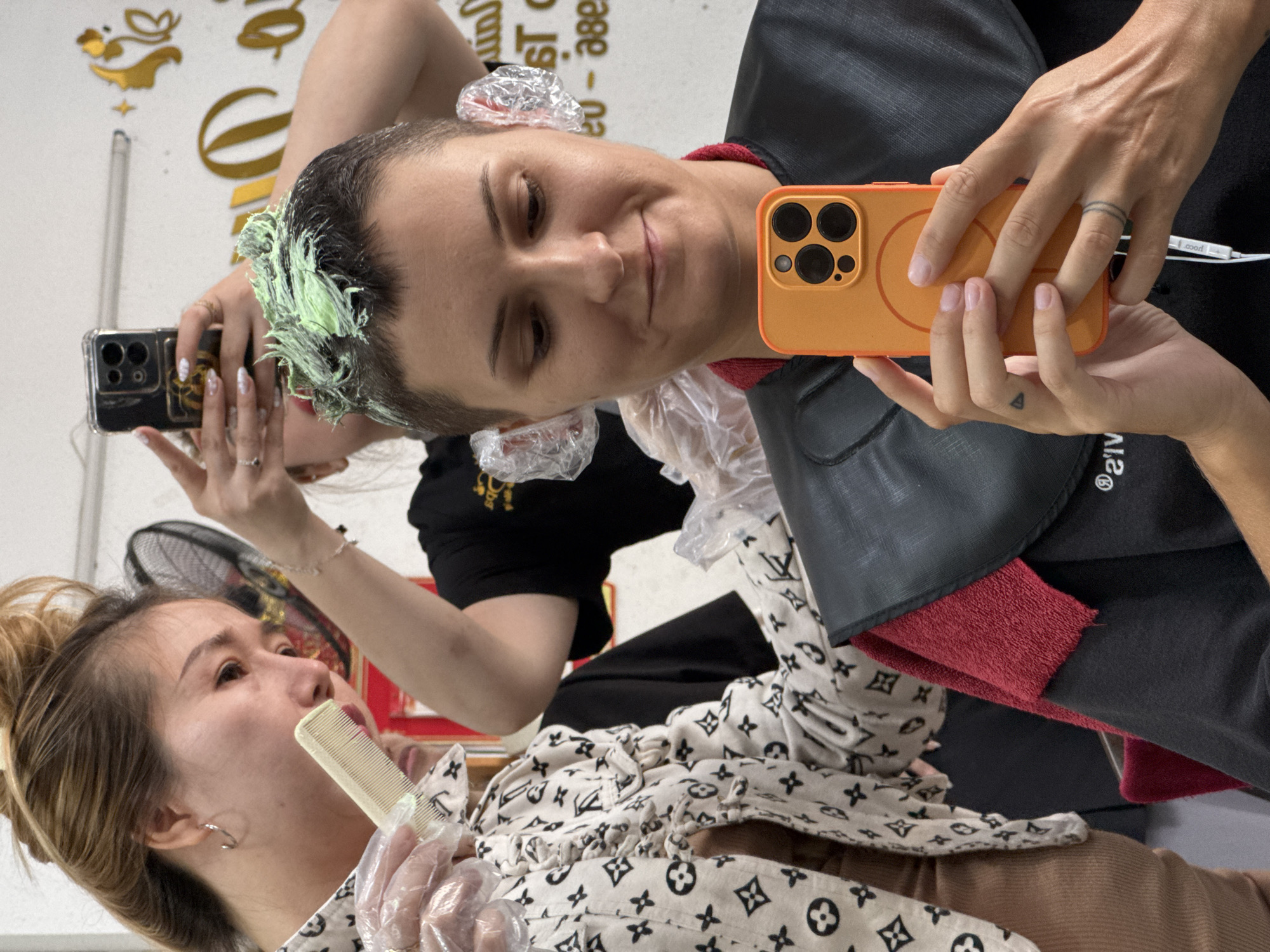 In a hair salon in Nha Trang Ward, Vietnam, a woman with short, dark hair is having her hair dyed a light green color. She is wearing a black smock over a red towel. She is holding an orange smartphone in her right hand and taking a selfie. Another woman, wearing gloves and a white top with a black pattern, is holding a comb and applying hair dye to the woman's hair. In the background, another person is taking a photo with a smartphone. The salon appears to be brightly lit.